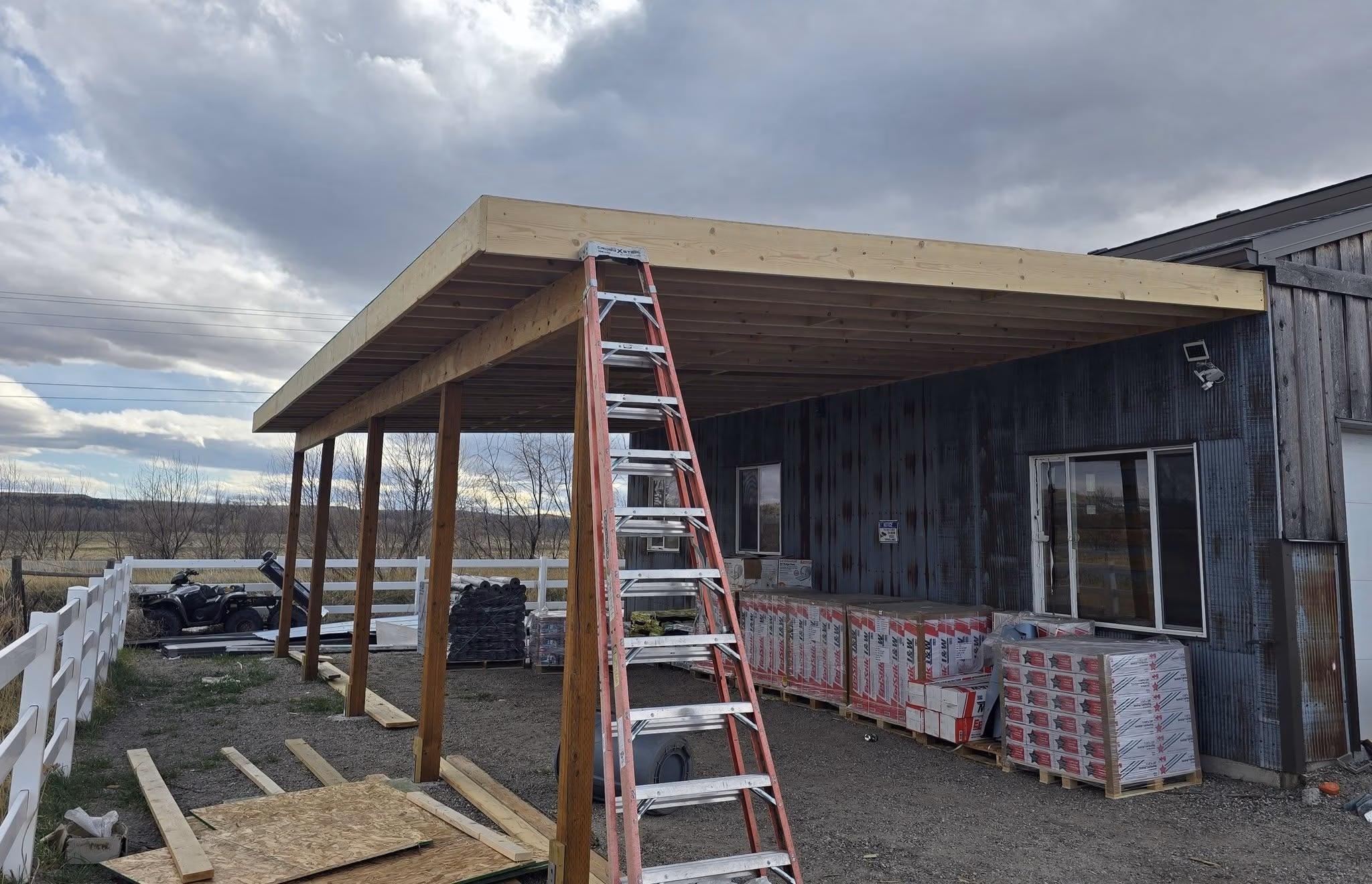 Wooden porch roof under construction with ladder, building materials, and cloudy sky.