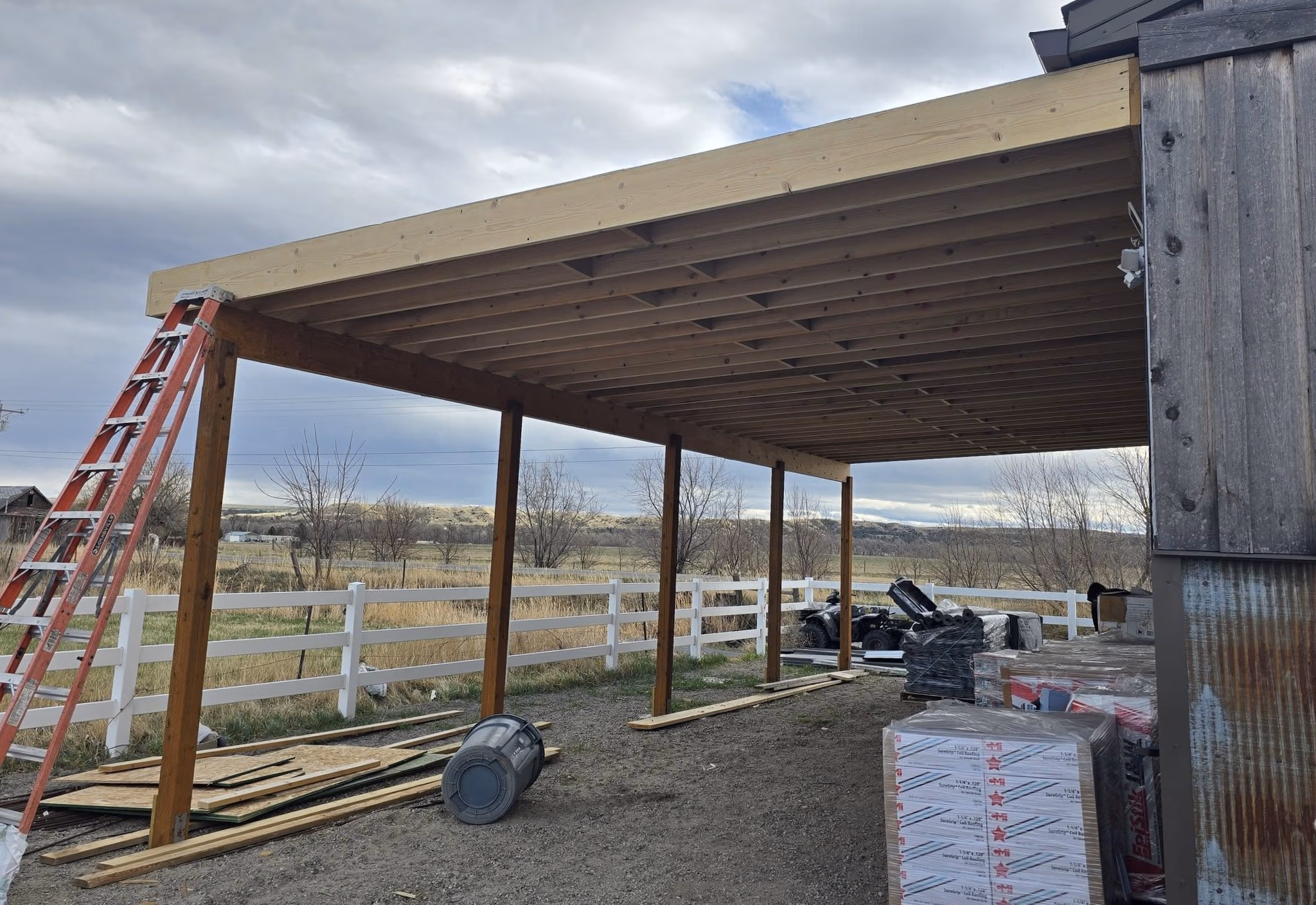 Under-construction wooden carport attached to a building with tools and materials around it.