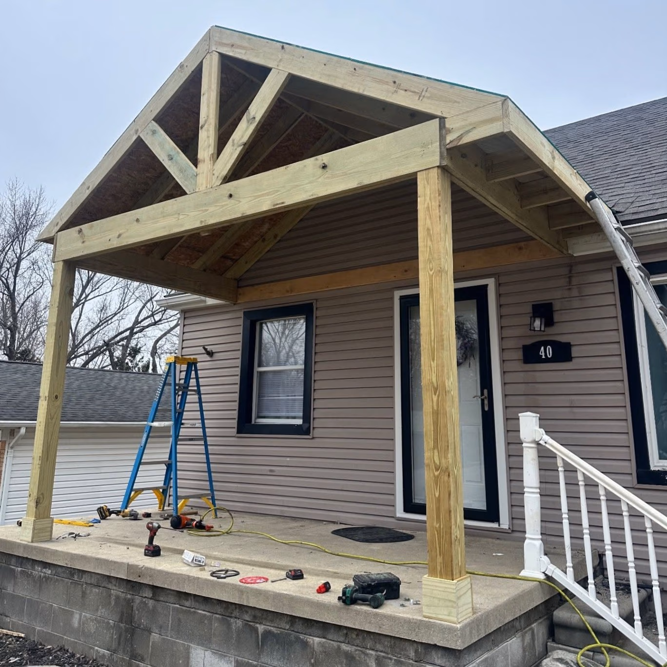 Partially built wooden porch roof structure in front of a house with tools and a ladder on the porch.