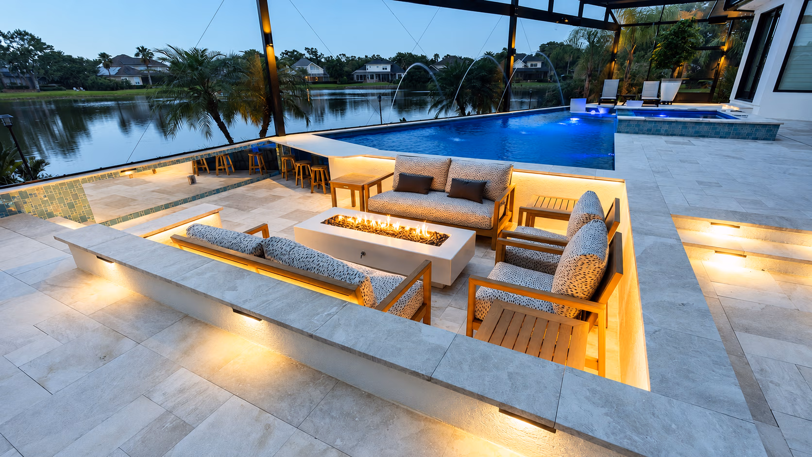 Modern patio with sunken seating area around firepit next to blue-lit pool overlooking a lake.