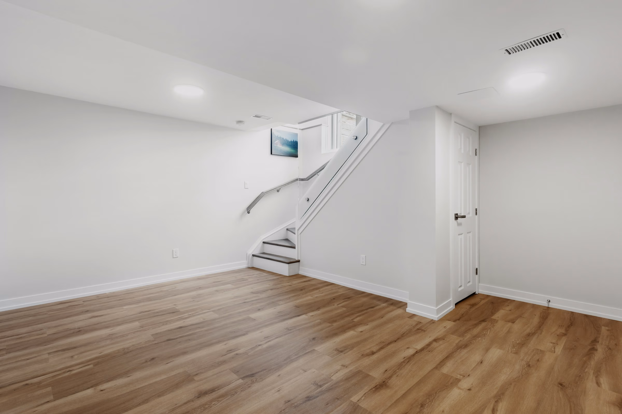 Bright basement room with wood flooring, white walls, stairs, and closed white door.