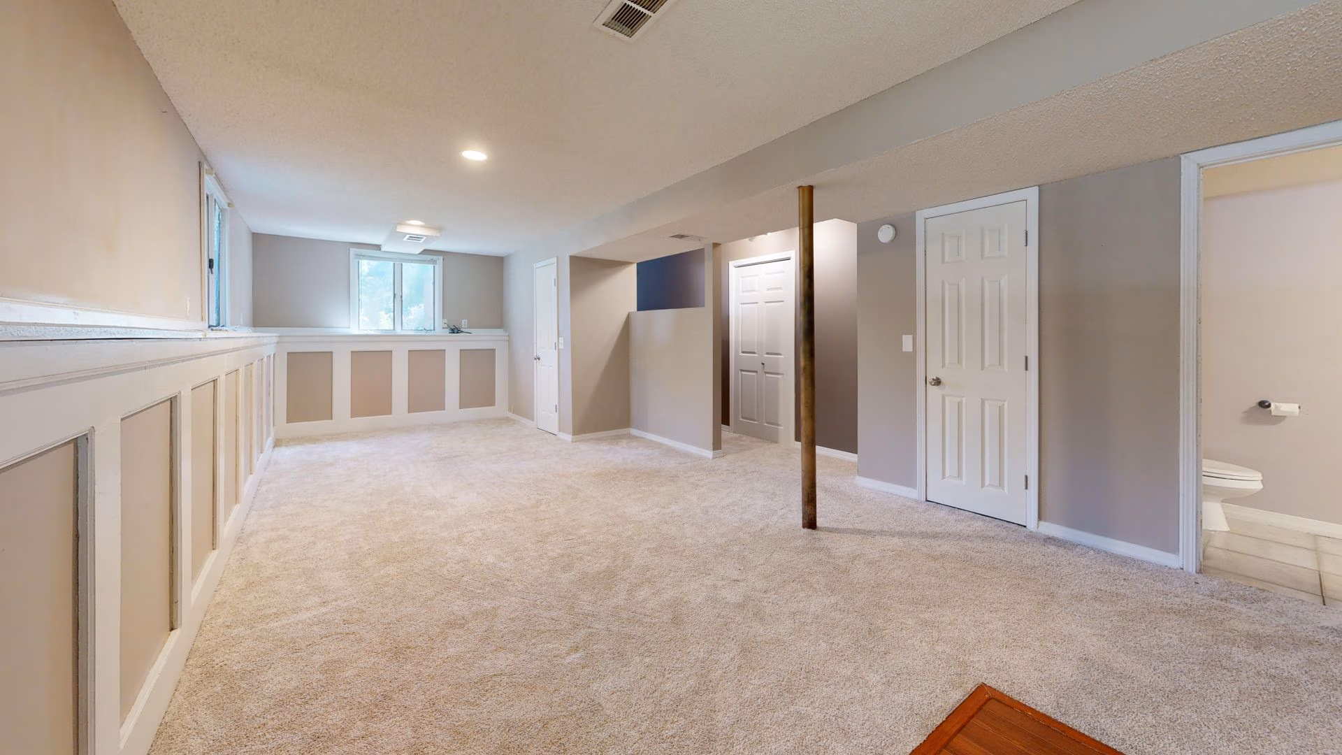 Empty finished basement room with beige carpet, wainscoting, white doors, and a small bathroom visible.