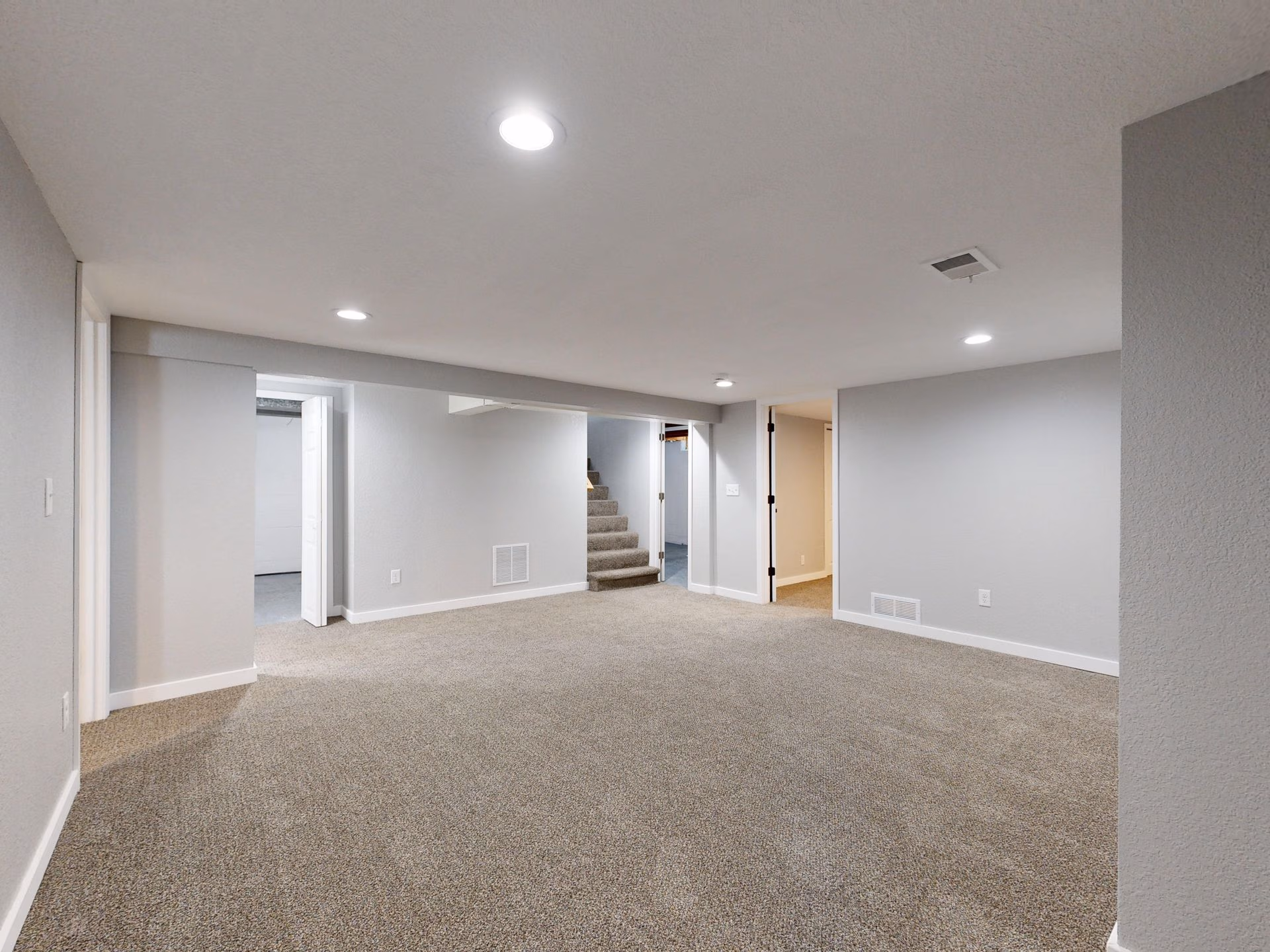 Spacious empty basement room with beige carpet, light gray walls, and recessed ceiling lights.