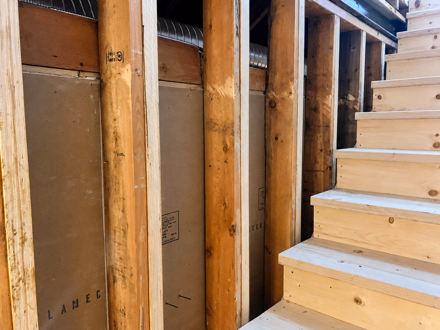 Wooden staircase beside unfinished basement wall framing with exposed wood studs and ductwork.