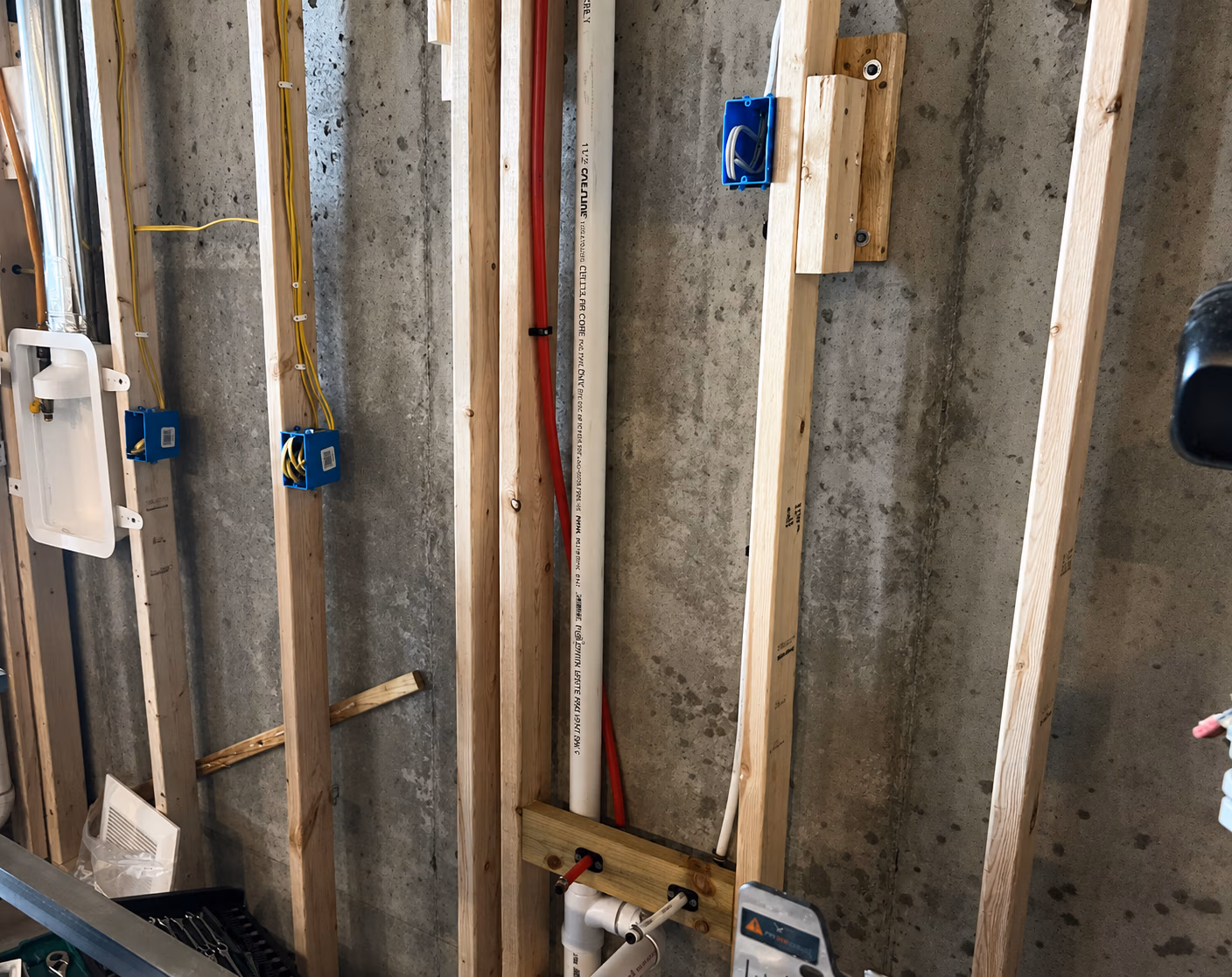 Basement wall with exposed wooden studs, electrical boxes, wiring, and plumbing pipes installed.