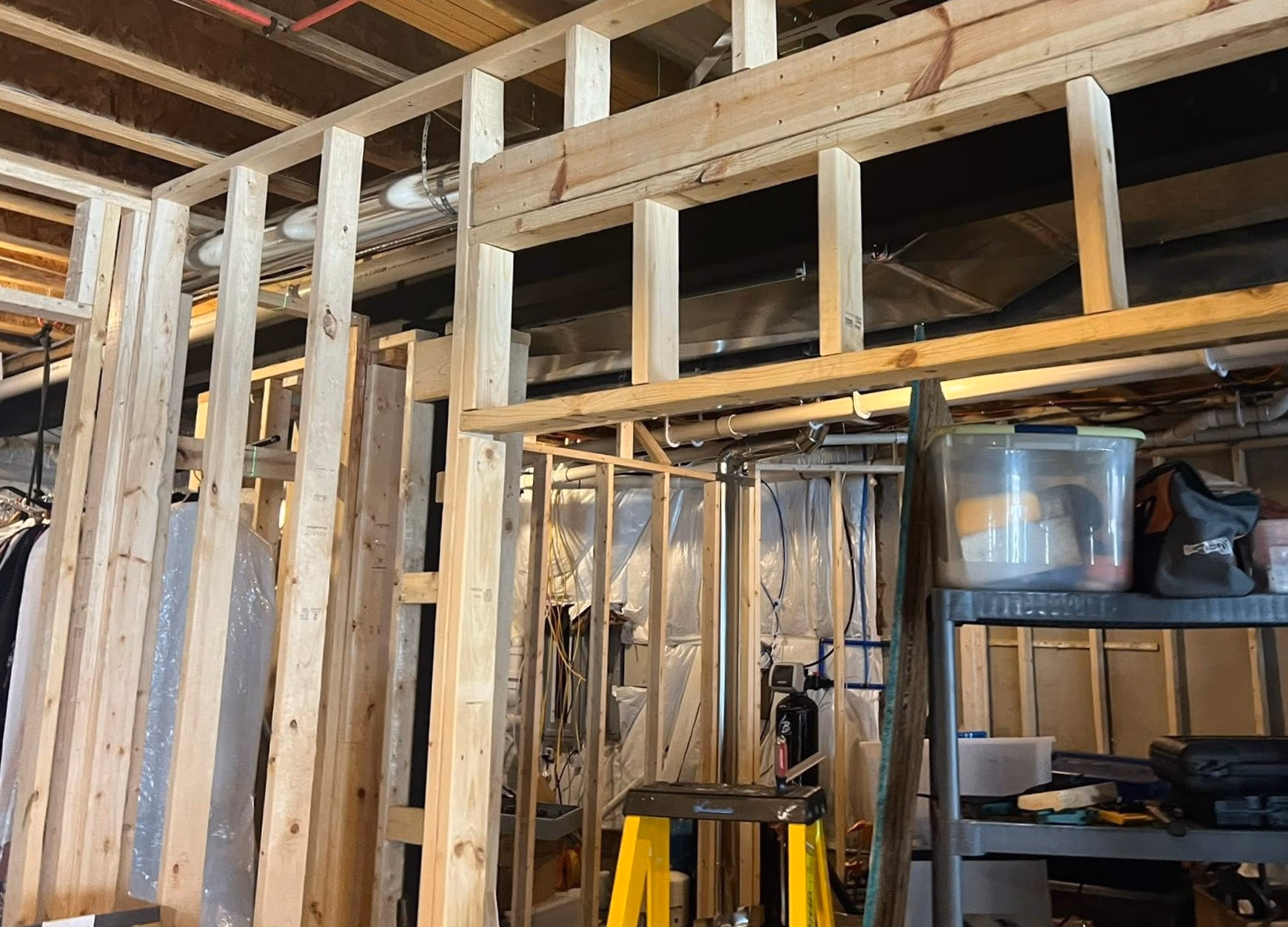 Basement under construction with exposed wooden studs, pipes, wiring, and a shelving unit with storage containers.
