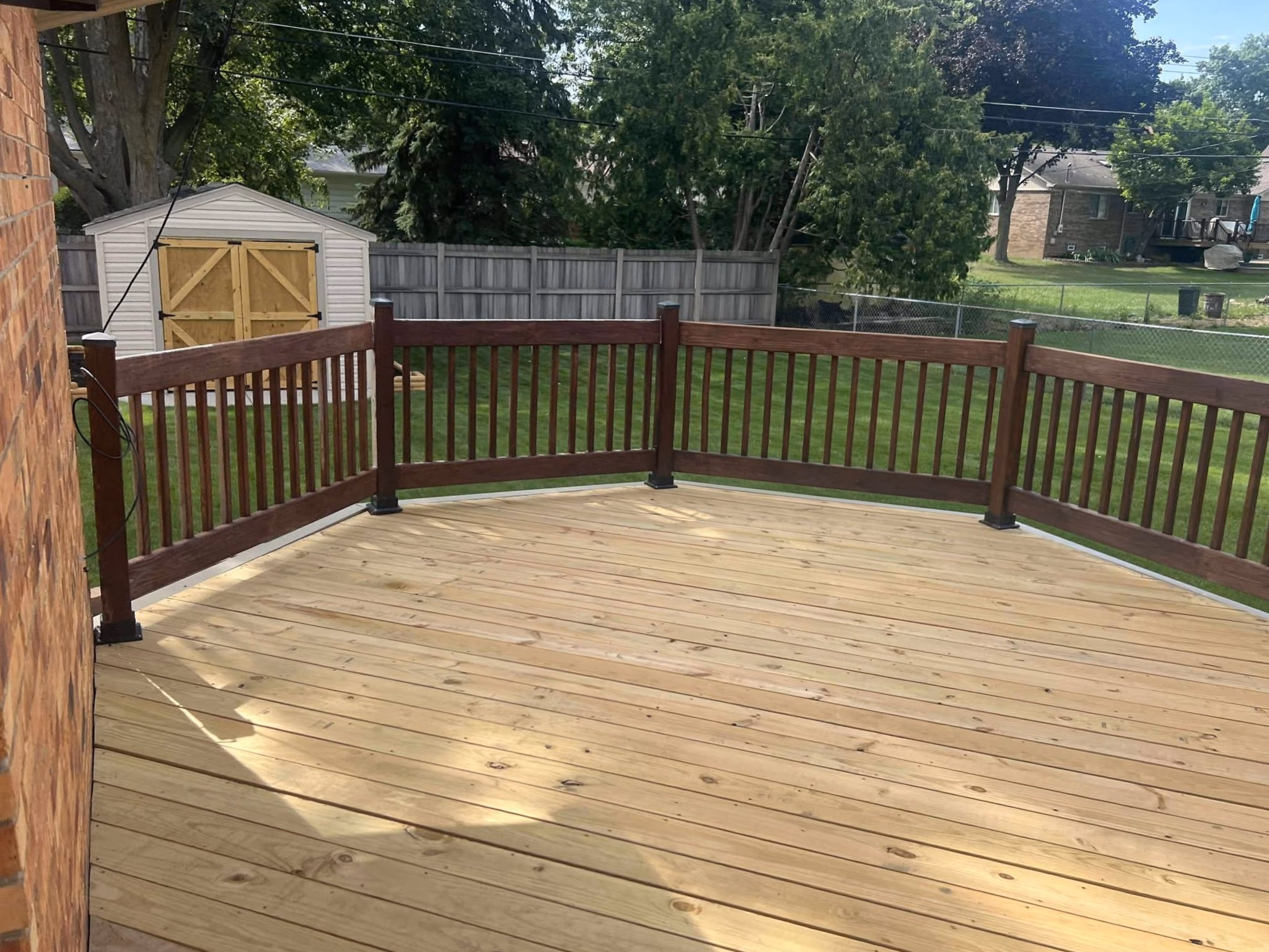 New wooden deck attached to a brick house with dark brown railing and green yard with a shed.