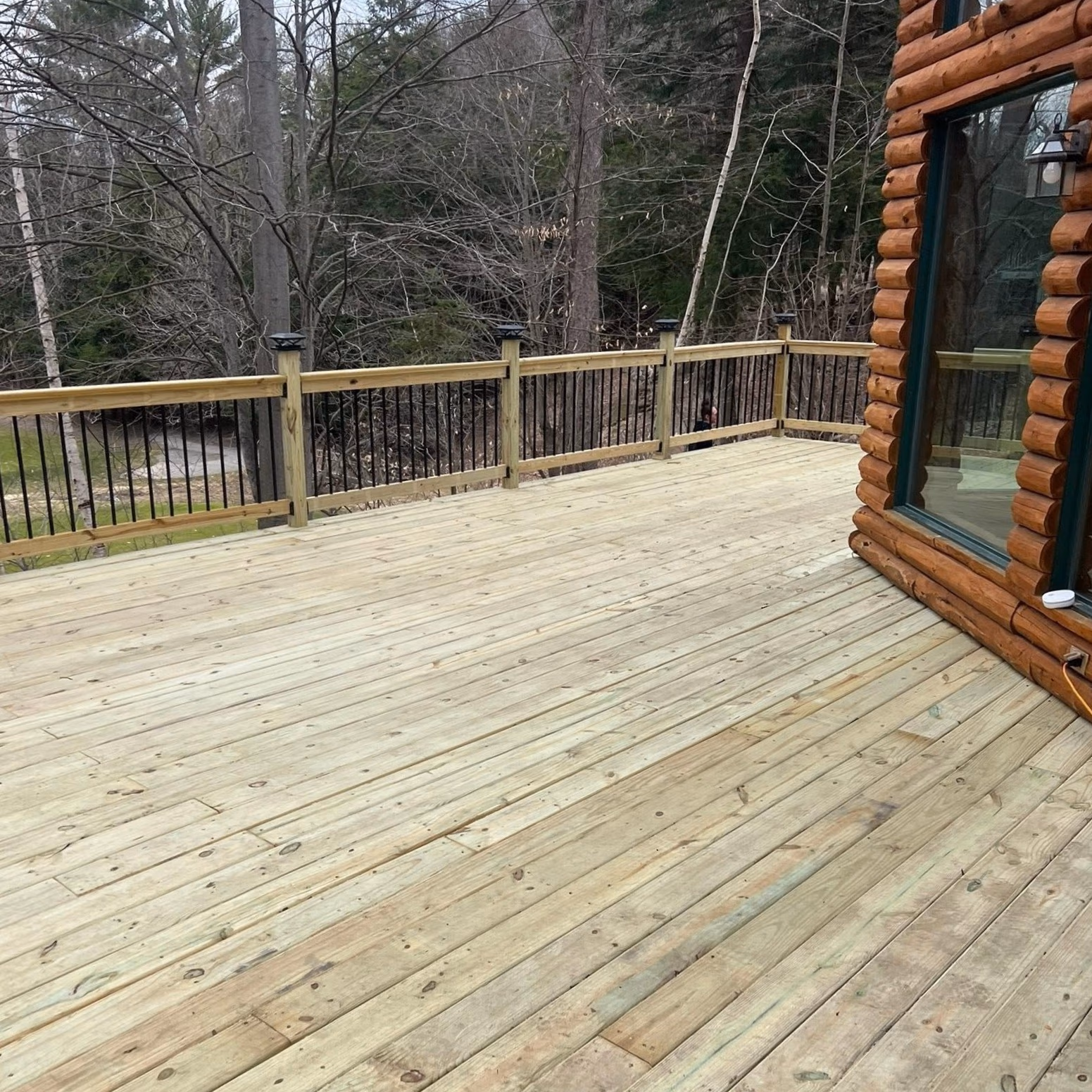 Spacious wooden deck attached to a log cabin, overlooking a forest with bare trees.