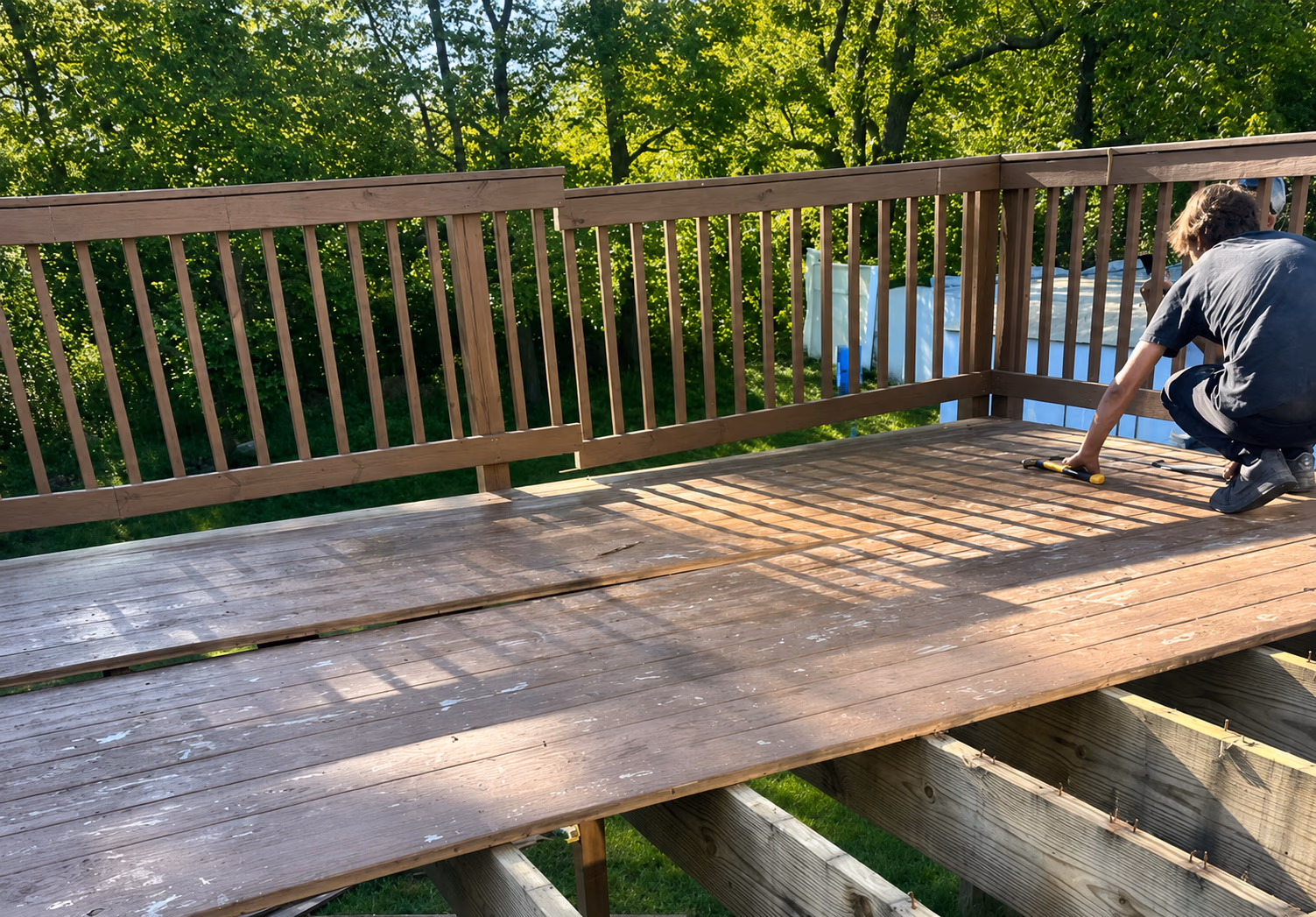 Person working on repairing a wooden deck with railing in a green backyard on a sunny day.