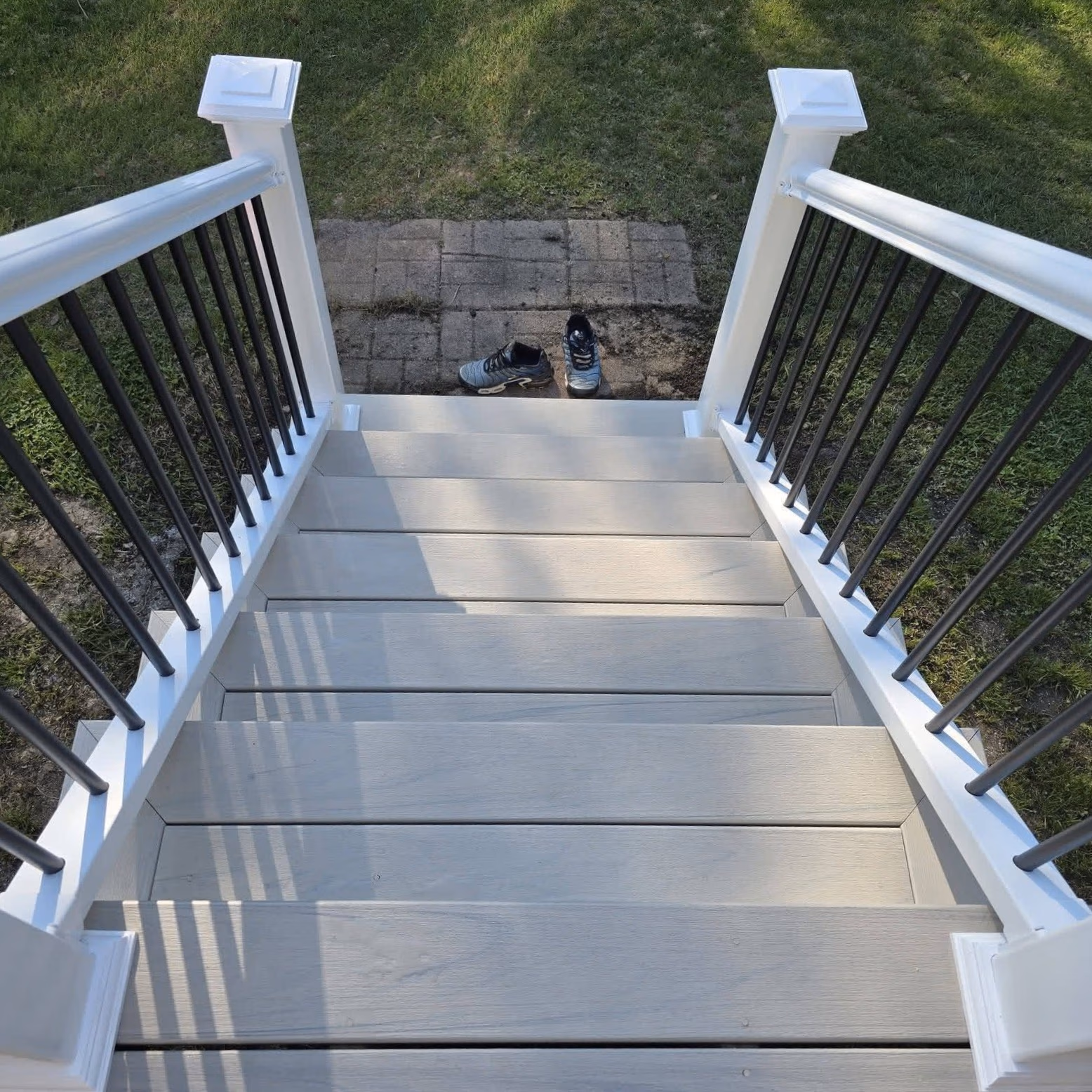 View down a gray wooden staircase with white posts and black balusters ending on a small dirt patio with two shoes.