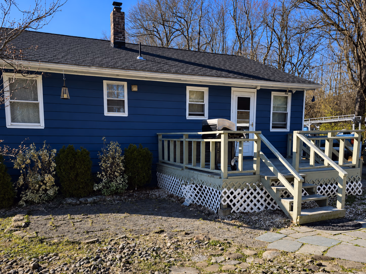 Blue house with a wooden deck, steps, a grill, and leafless trees in the background.