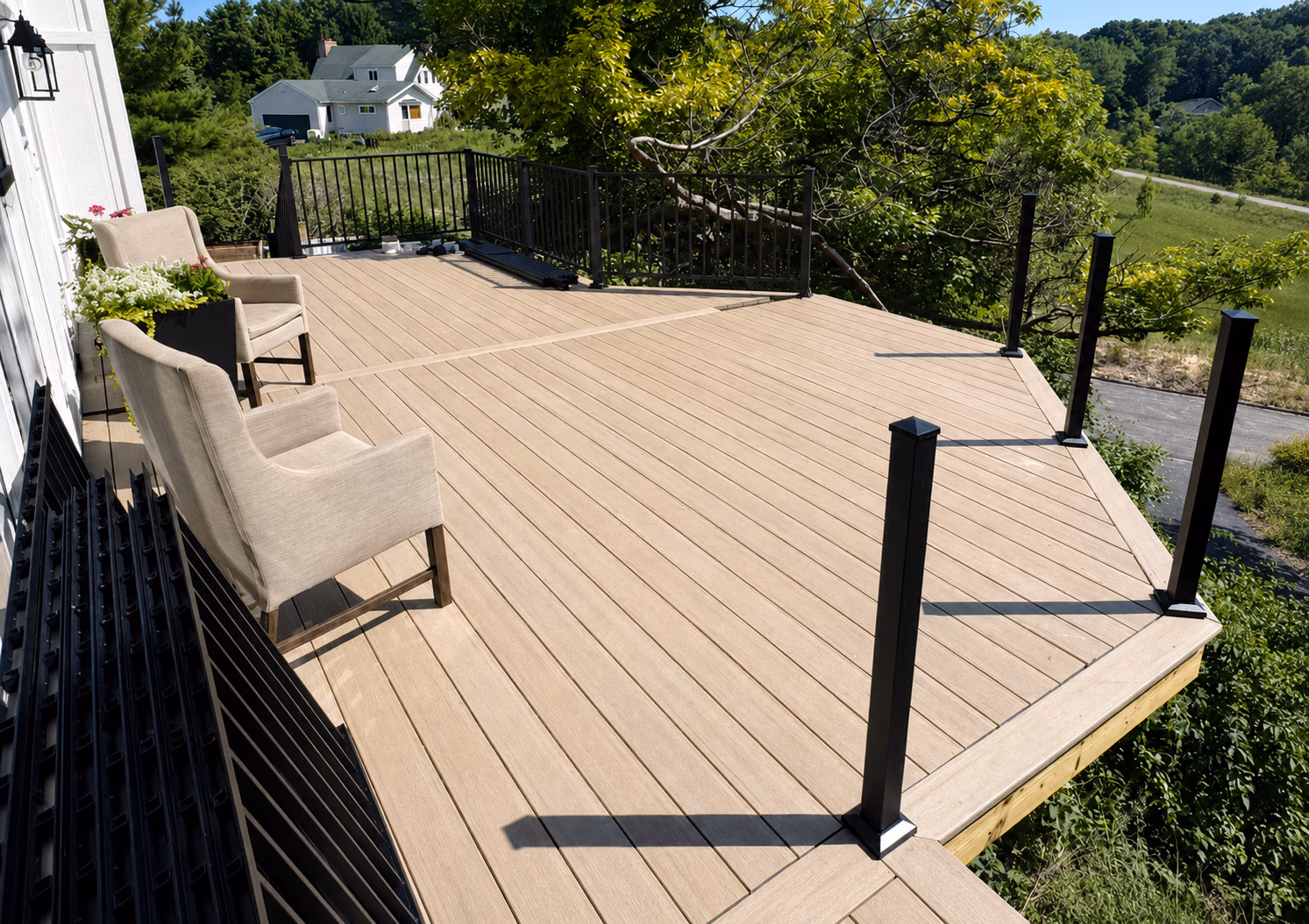 Spacious wooden deck with black railing posts, two beige chairs, and green plants outdoors.
