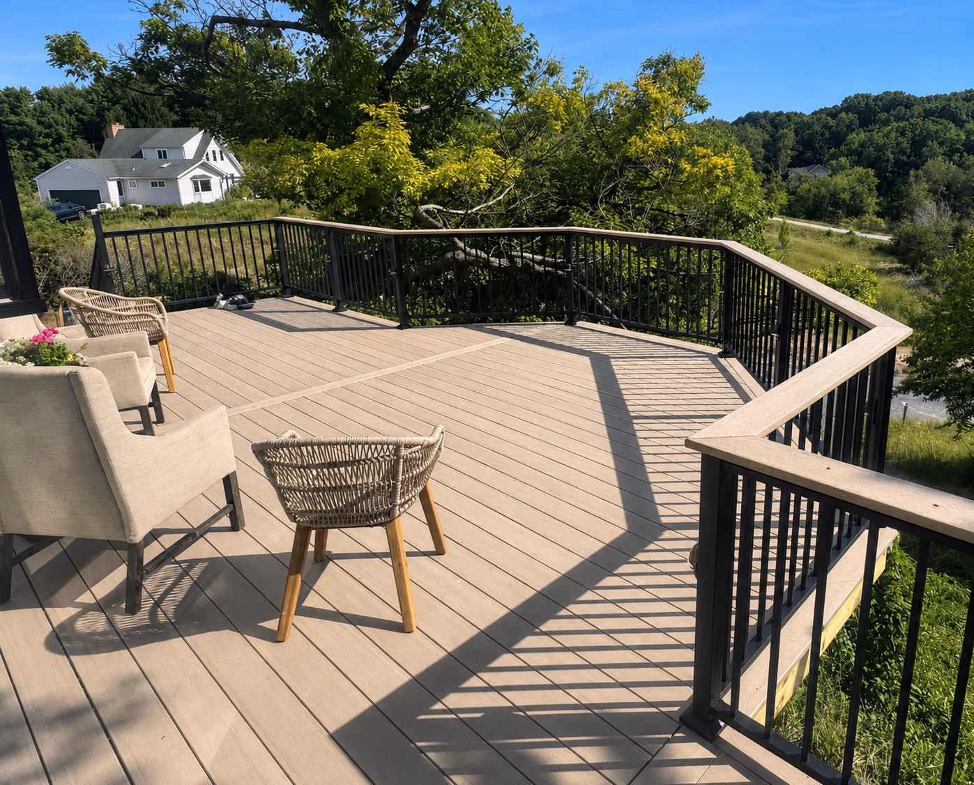 Spacious wooden deck with black railing, two beige chairs, a woven chair, overlooking green trees and a house.