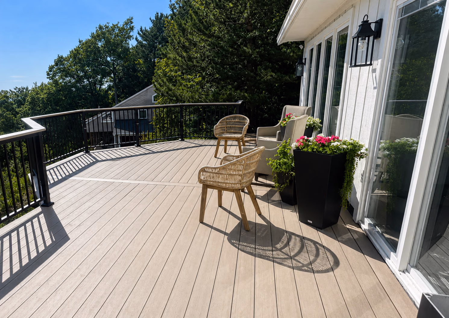 Spacious outdoor deck with wicker chairs, armchairs, flower pots, and a black railing under a clear sky.