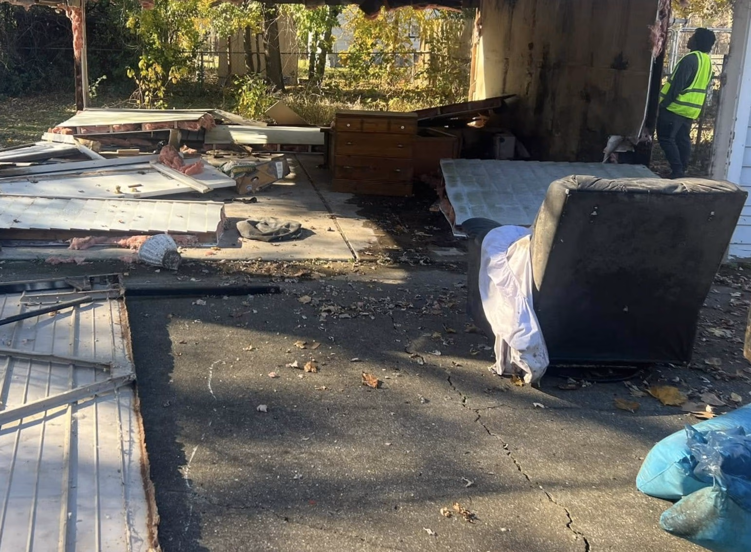 Person in safety vest stands near a damaged garage with scattered debris and furniture outside.