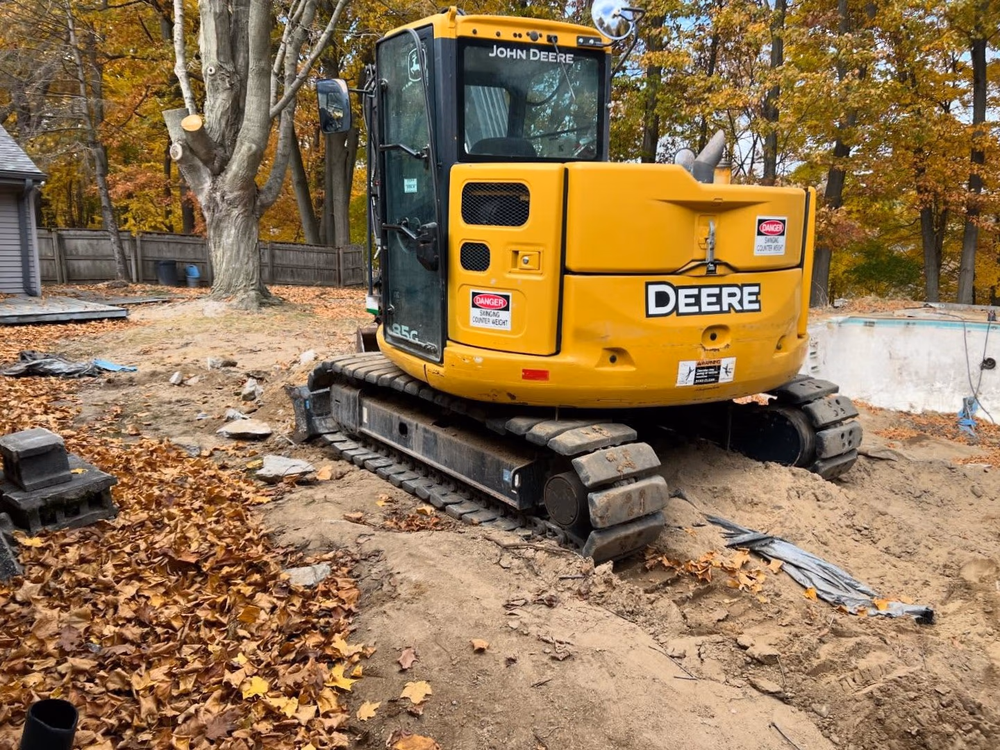 Yellow John Deere excavator on dirt surrounded by autumn trees and fallen leaves near a drained pool.