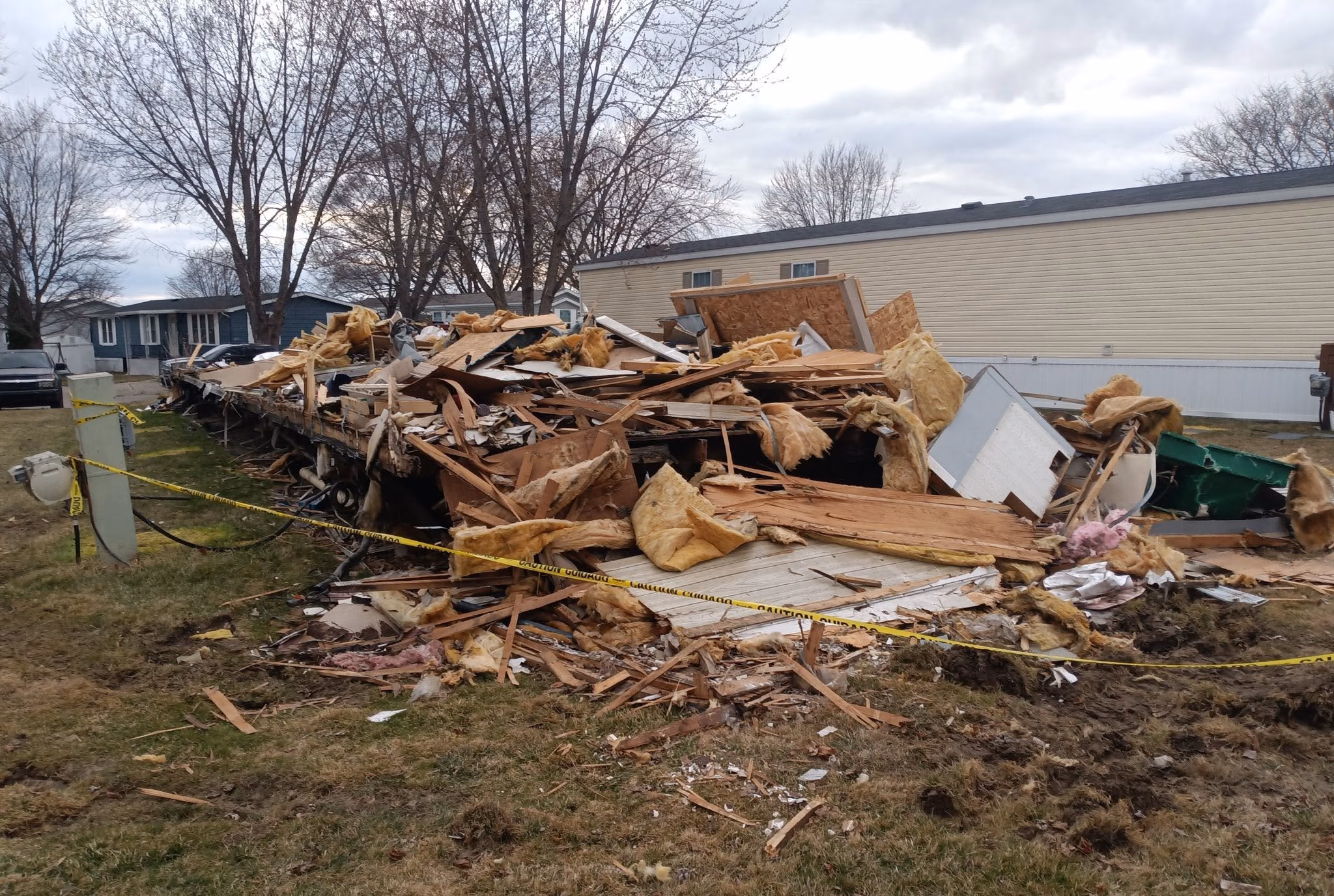 Demolished mobile home remains with insulation and debris piled on grass, marked by caution tape.