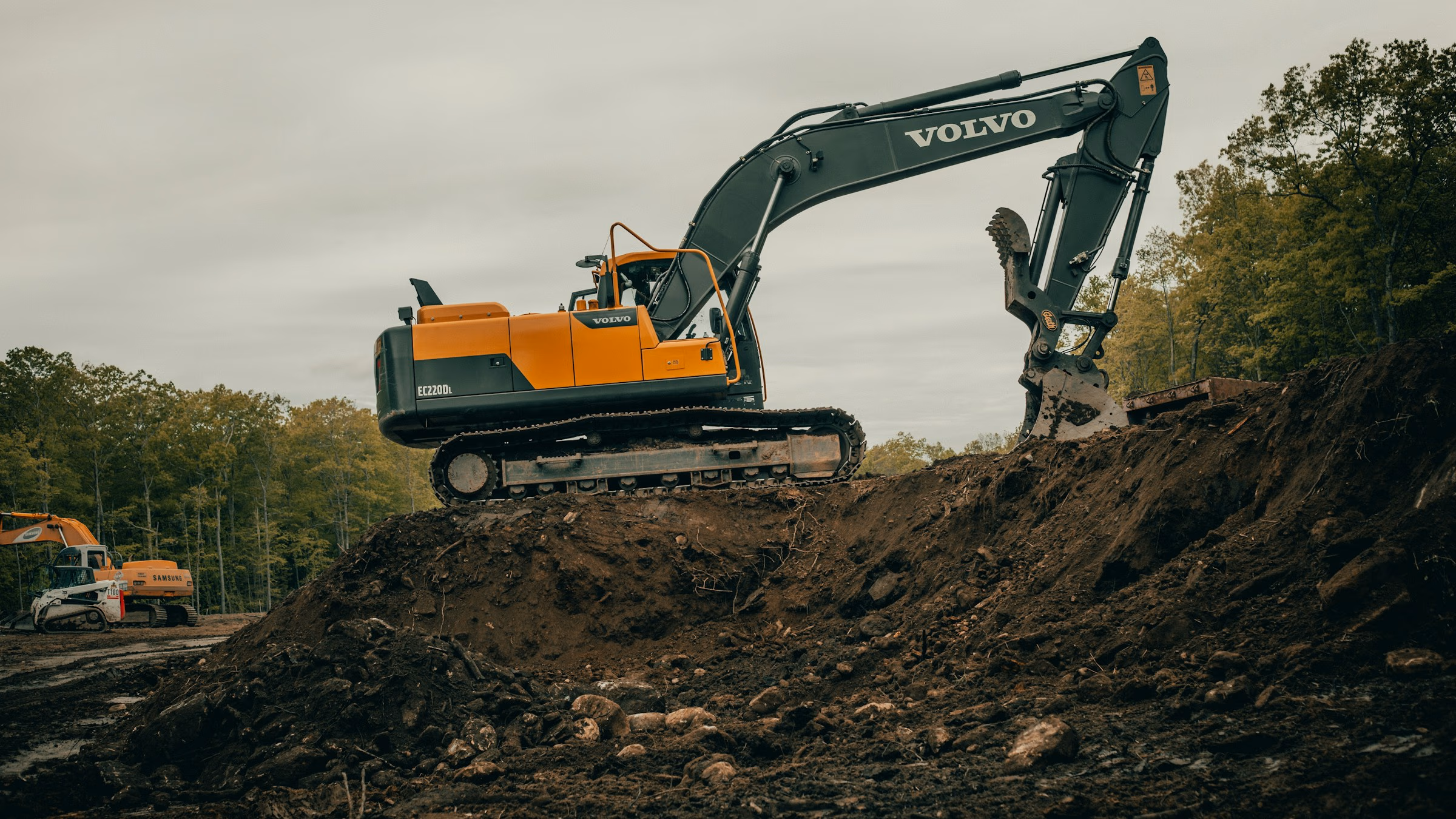 Yellow Volvo EC220DL excavator digging dirt on construction site with trees in background.