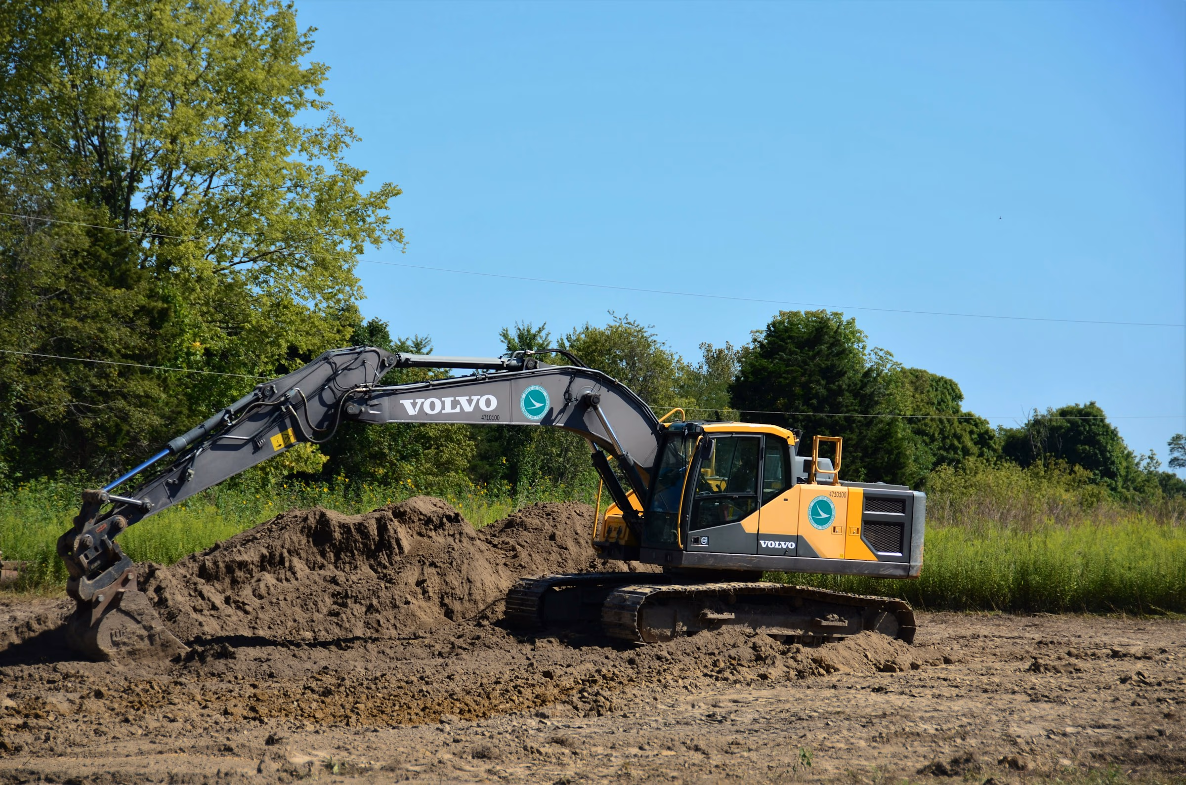 Yellow and gray Volvo excavator digging and moving dirt in a green, tree-lined field under blue sky.