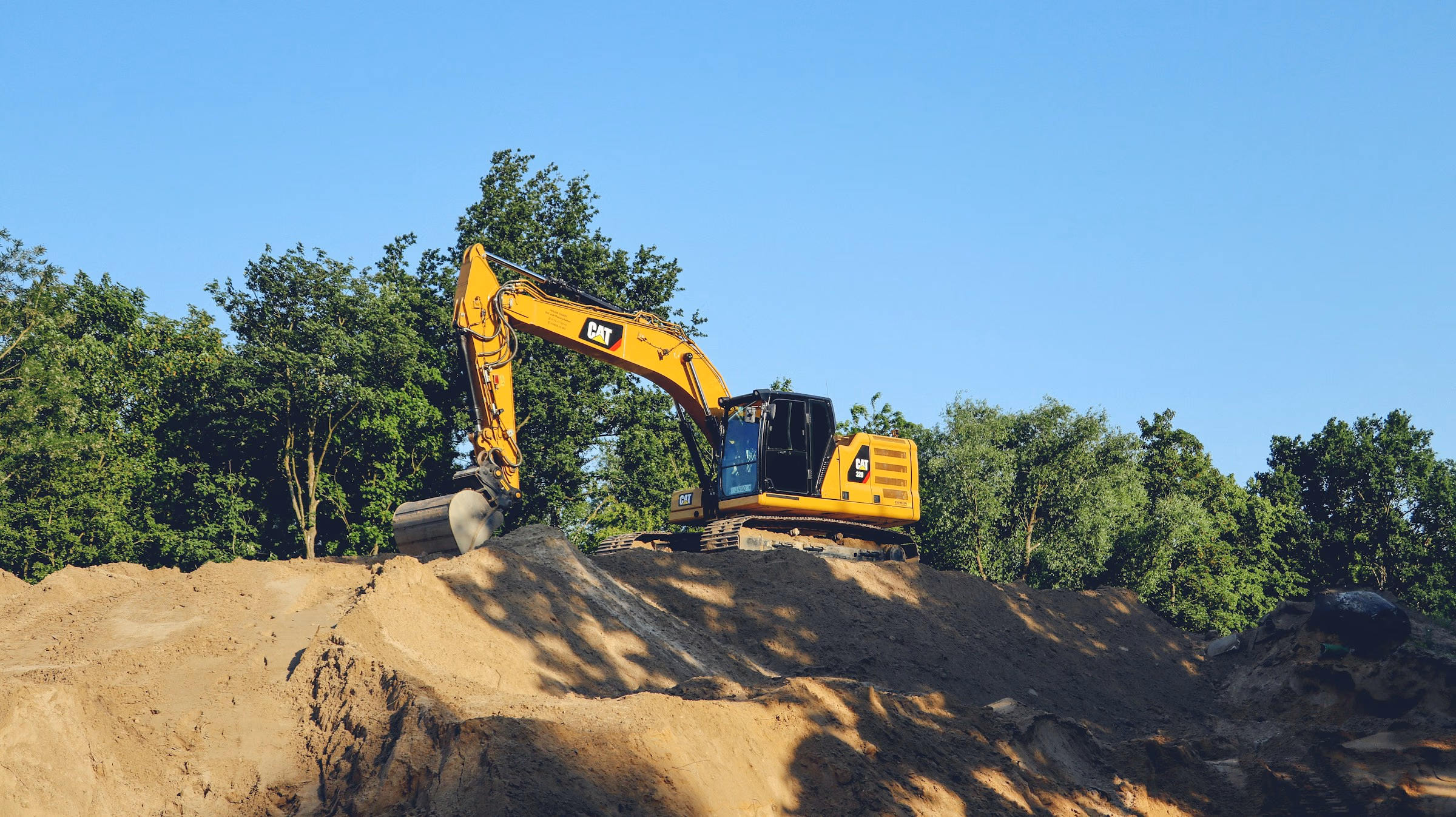 Yellow CAT excavator on dirt mound with green trees and clear blue sky in background.