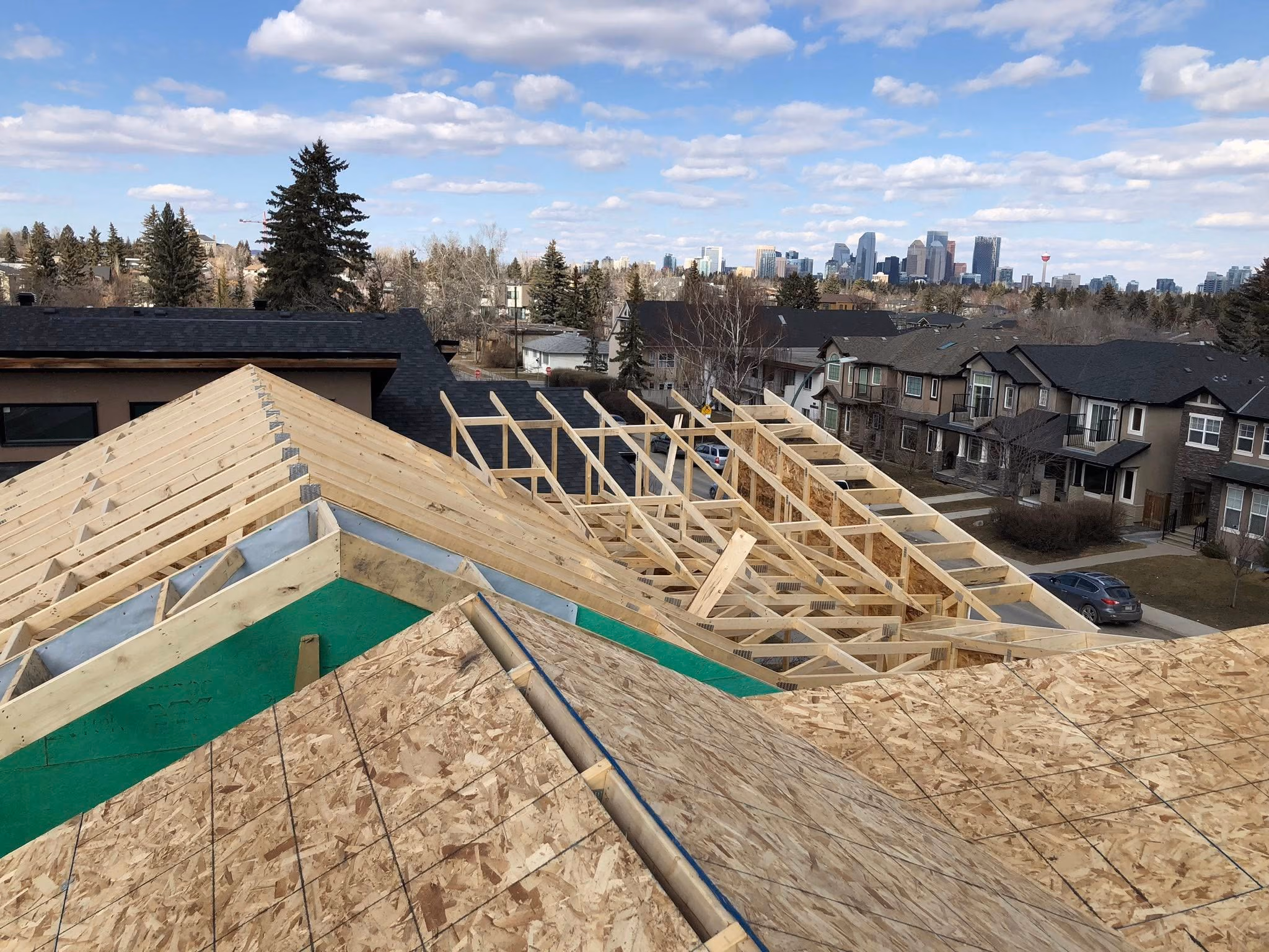 Roof under construction with wooden framing and sheathing, suburban houses, and city skyline in the background.