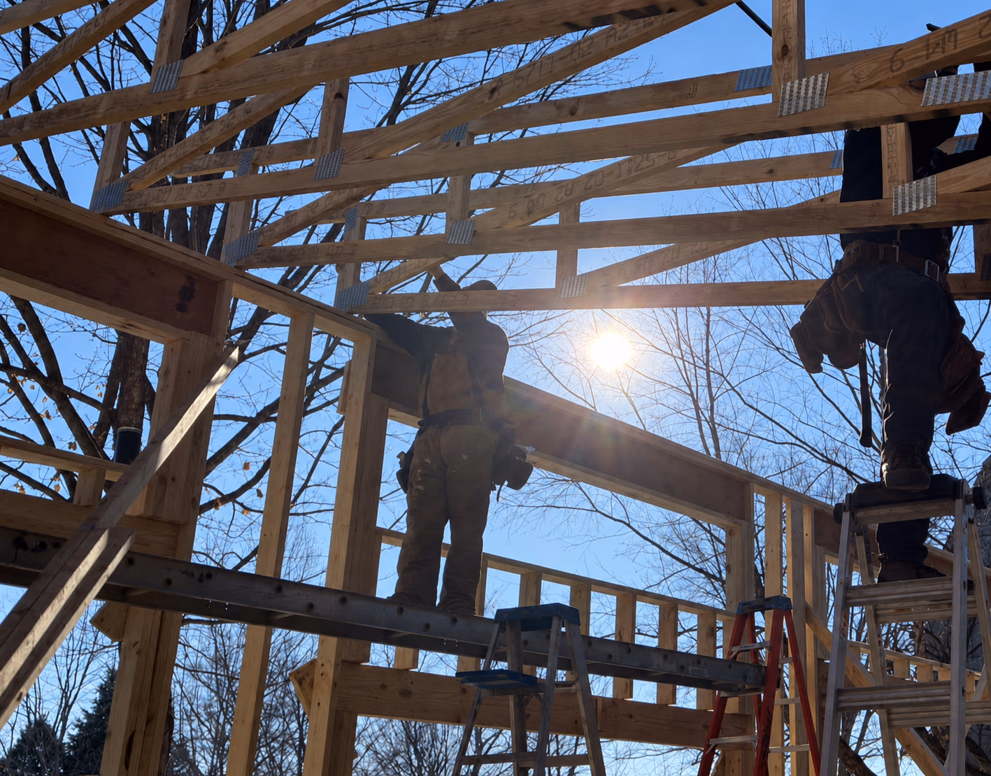 Two construction workers framing a wooden structure with ladders under bright sunlight and blue sky.