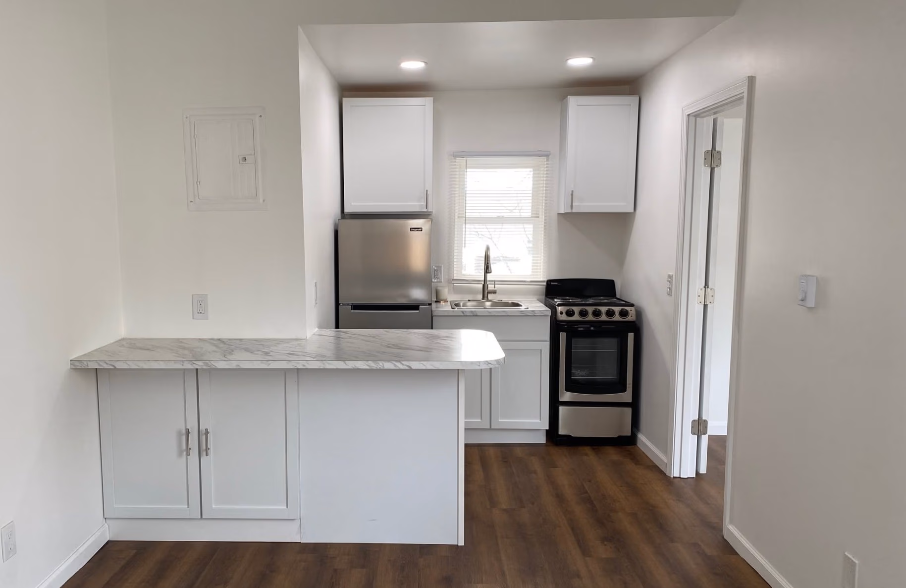 Small modern kitchen with white cabinets, marble countertop, stainless fridge, stove, and wooden floor.