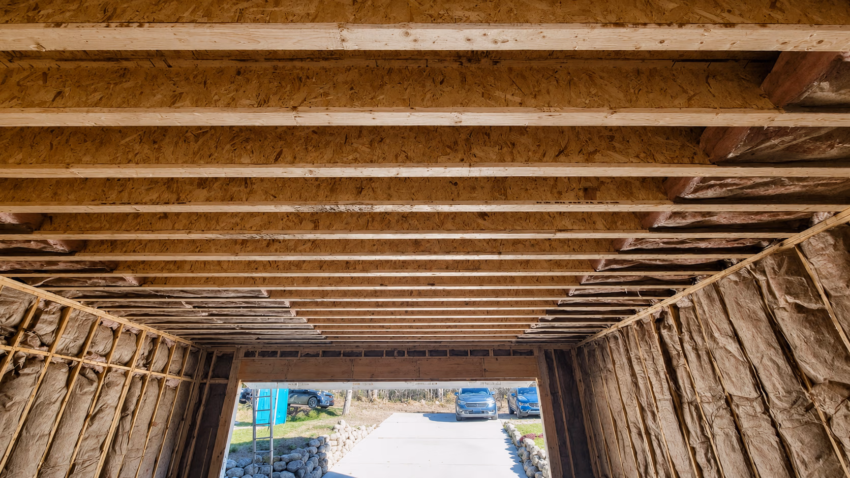 Interior of a wooden framed garage under construction with insulation on walls and cars outside.