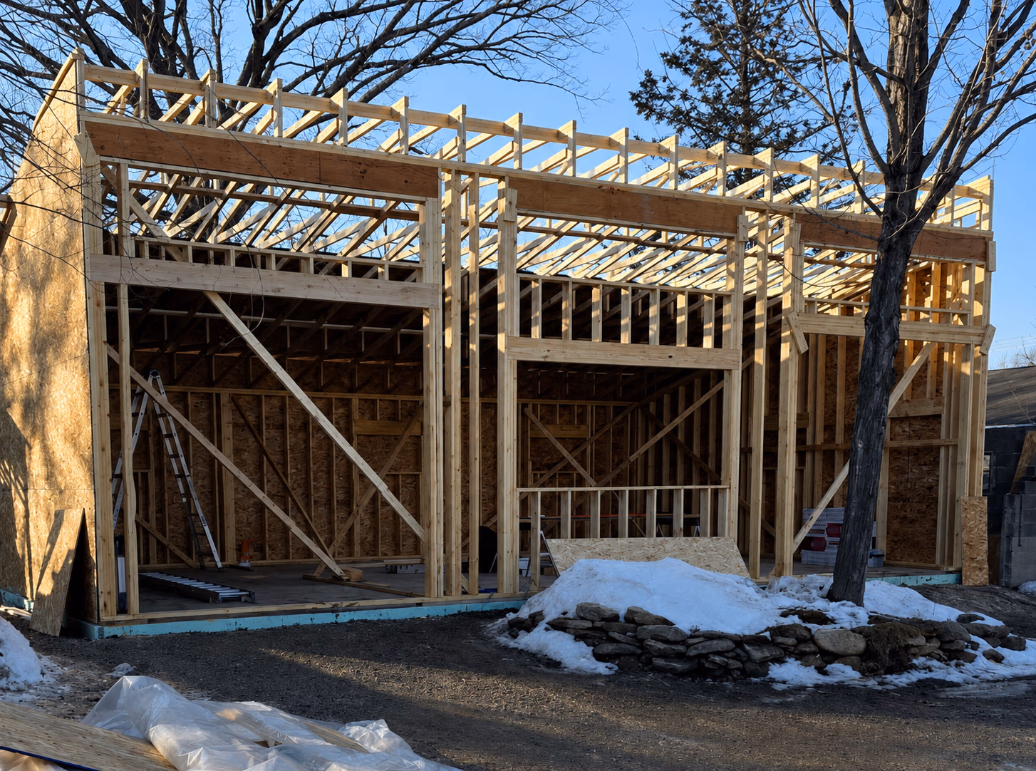 Wooden frame structure of a building under construction with some snow and trees nearby.