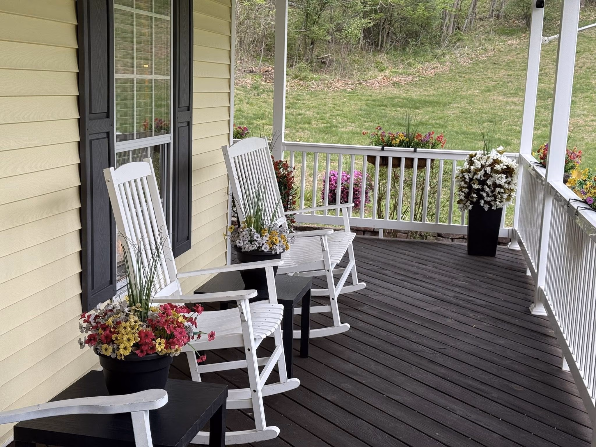 Front porch with white rocking chairs, black tables, potted flowers, and yellow siding walls.