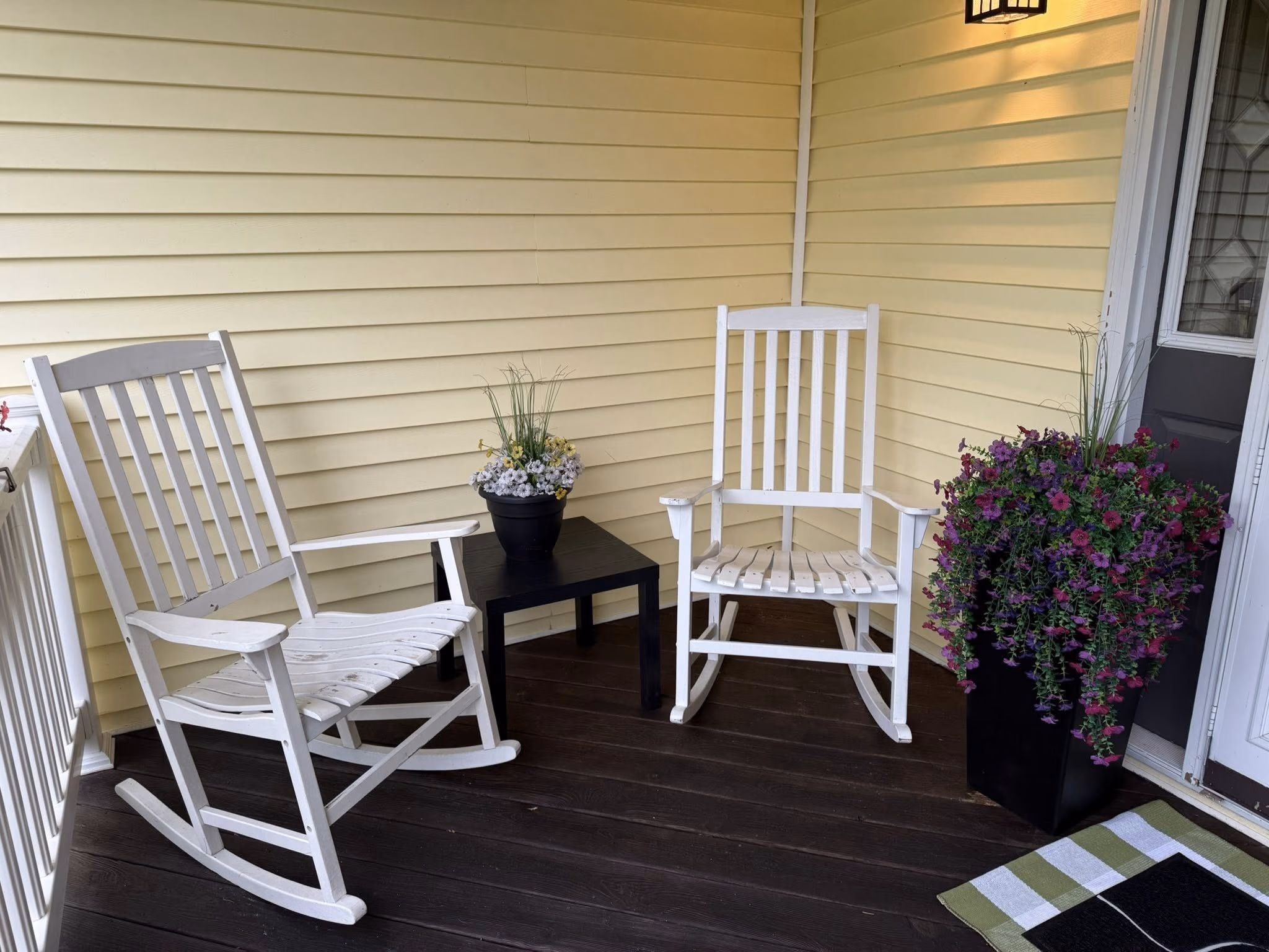 Two white rocking chairs on dark wood porch with black side table and flower pots near yellow siding.