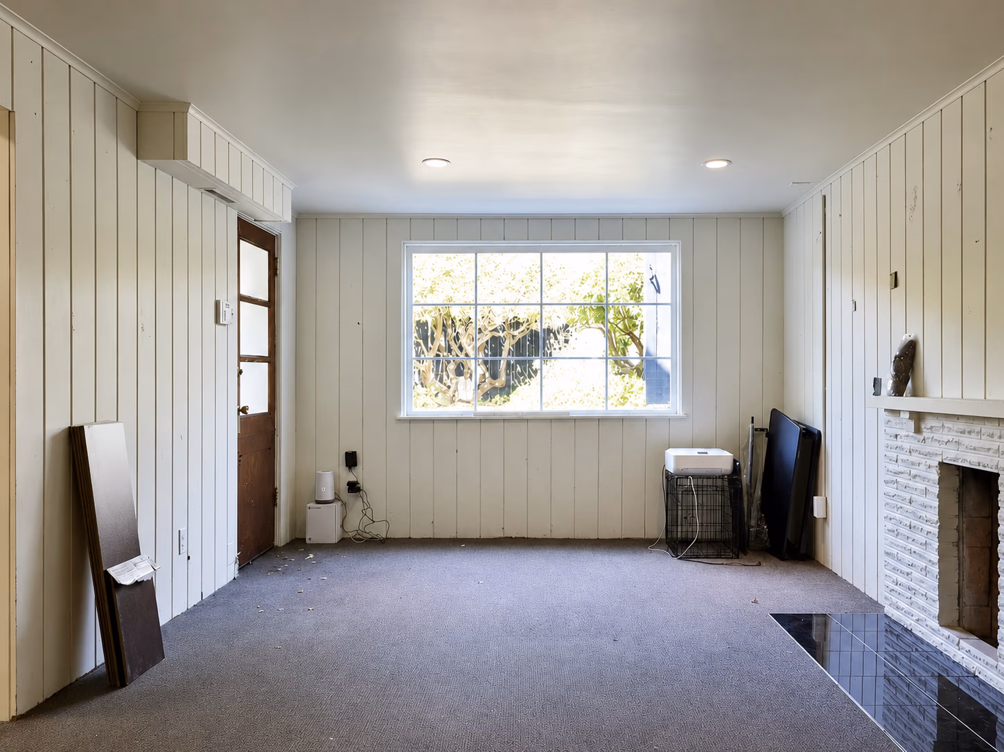 Empty room with white paneled walls, large window, carpeted floor, and a fireplace on the right.