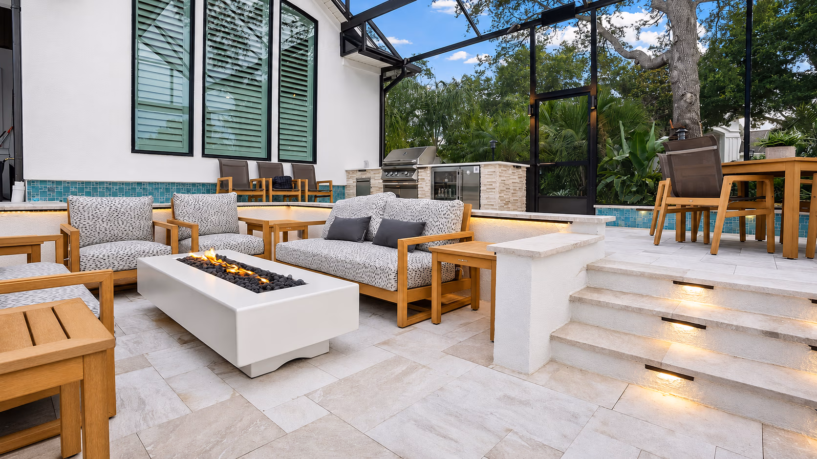 Modern outdoor patio with cushioned wooden furniture, fire pit, built-in grill, and tiled flooring.