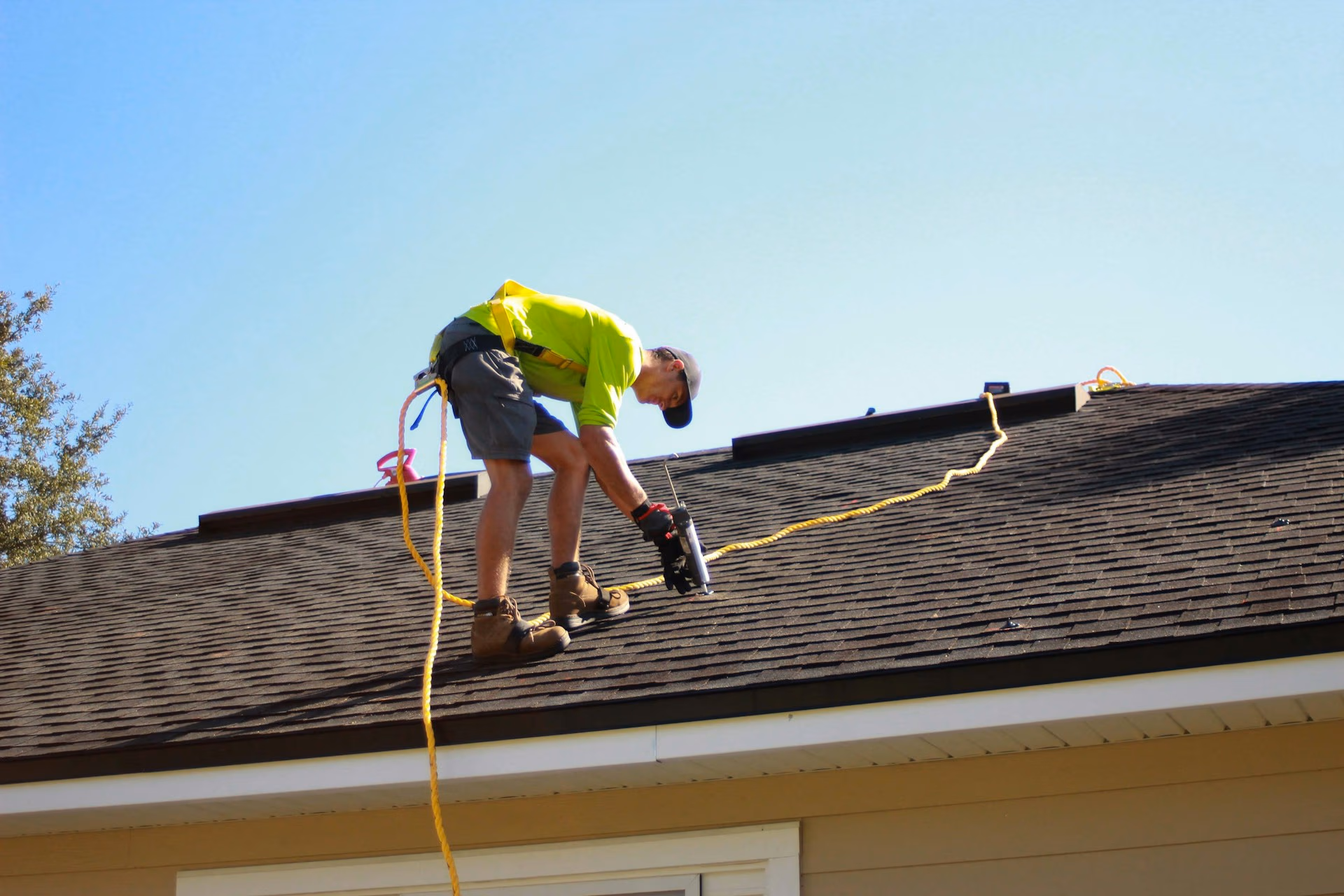 Worker in safety gear repairing a shingled roof under clear blue sky.