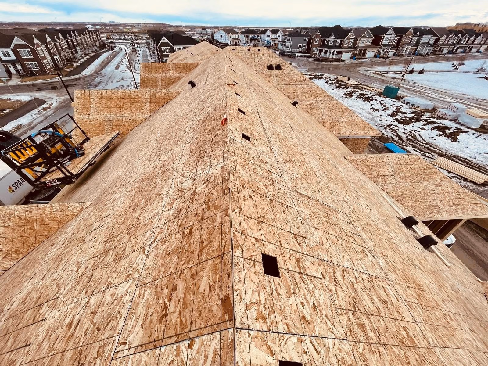 View of a large wooden roof under construction on a residential building with houses and snow nearby.