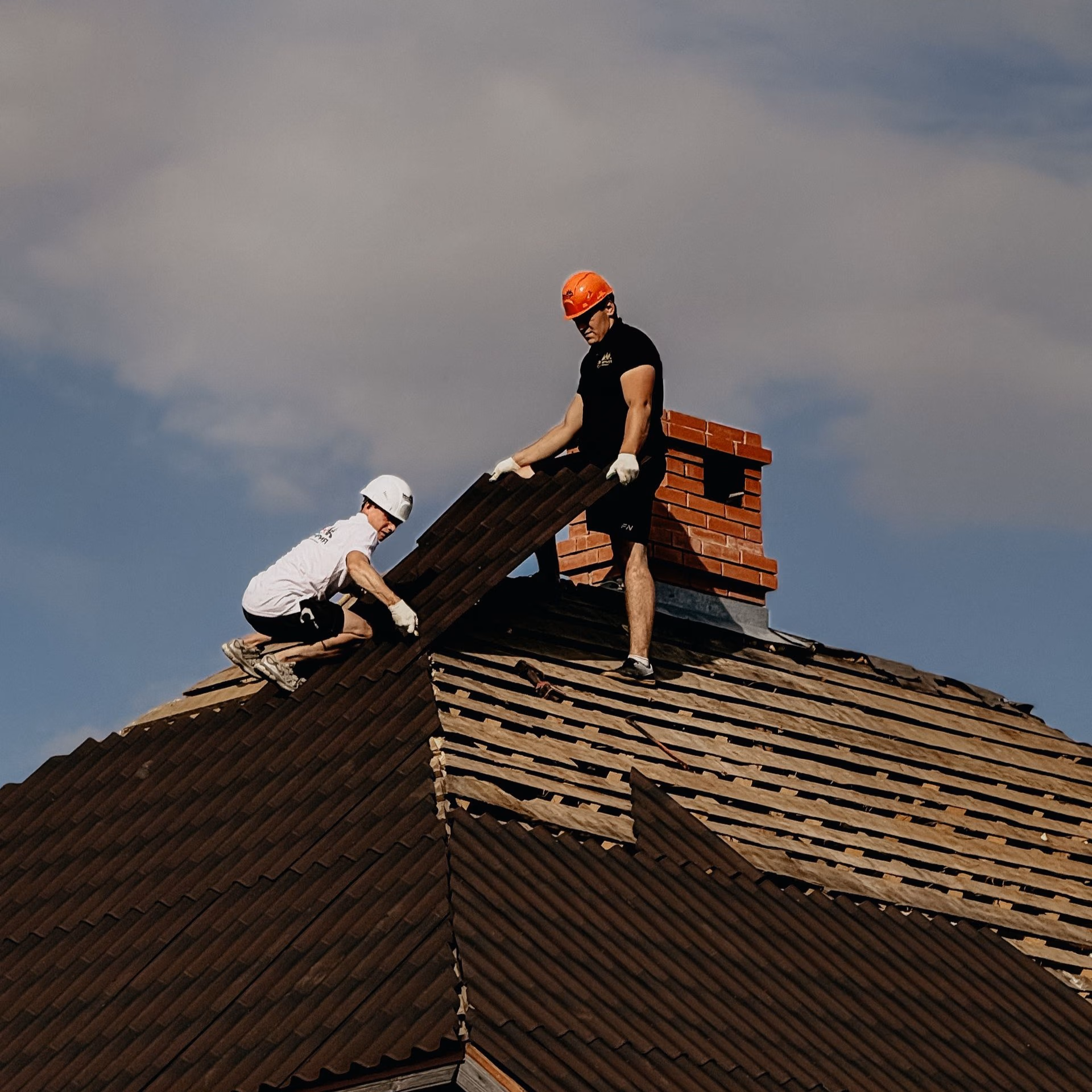 Two workers wearing helmets installing brown shingles on a rooftop with a brick chimney.