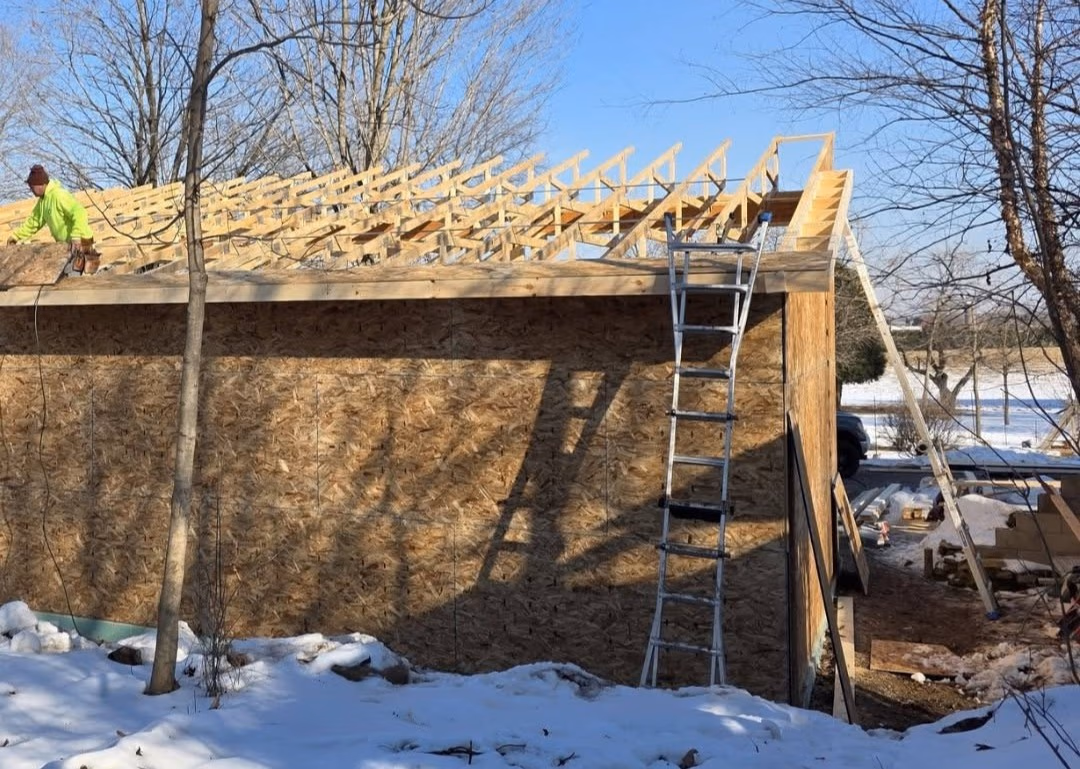 Construction worker building roof frame on a wooden structure in a snowy yard with ladders nearby.