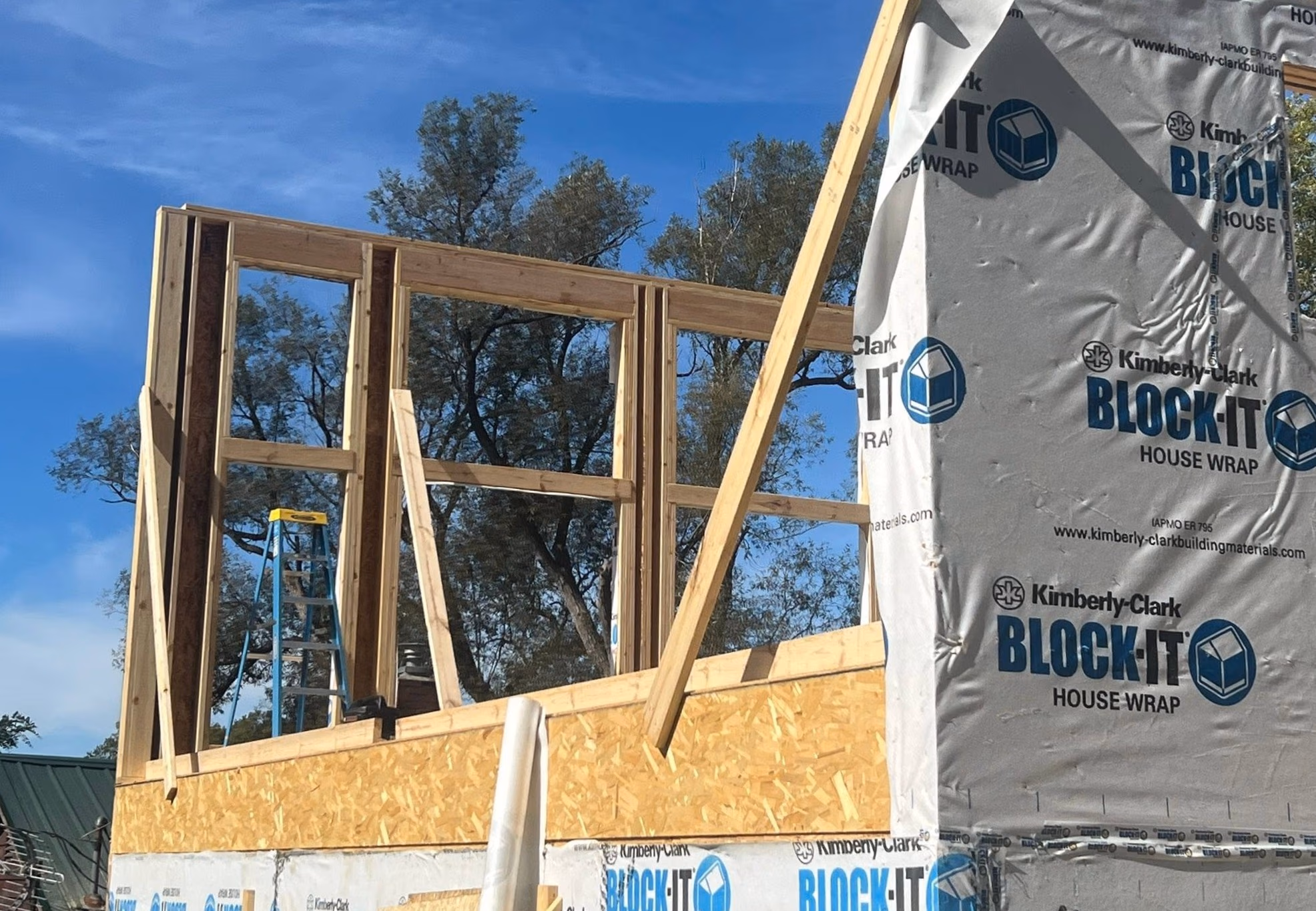 Wooden framing and house wrap on a building under construction with trees and blue sky in background.