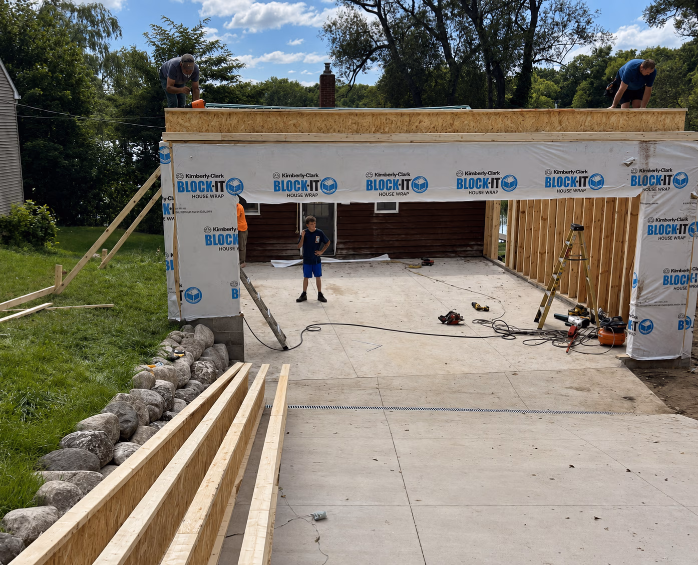 Construction site with workers building structure covered in Block-It house wrap on sunny day.