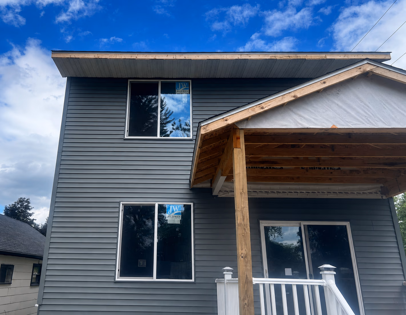 Two-story house with gray vinyl siding, two double-pane windows, a porch under construction, and blue sky.