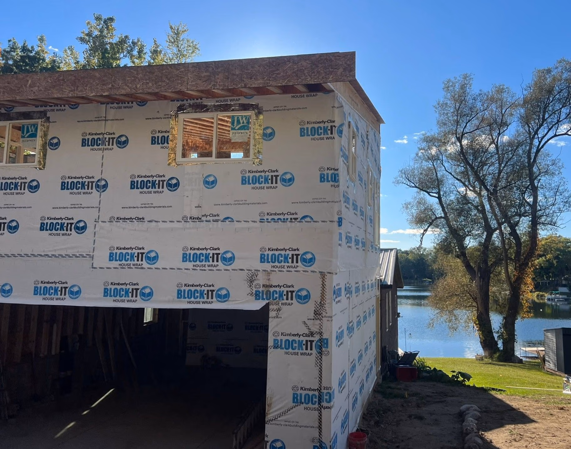 Unfinished two-story building covered in house wrap near a lake with trees and clear blue sky.