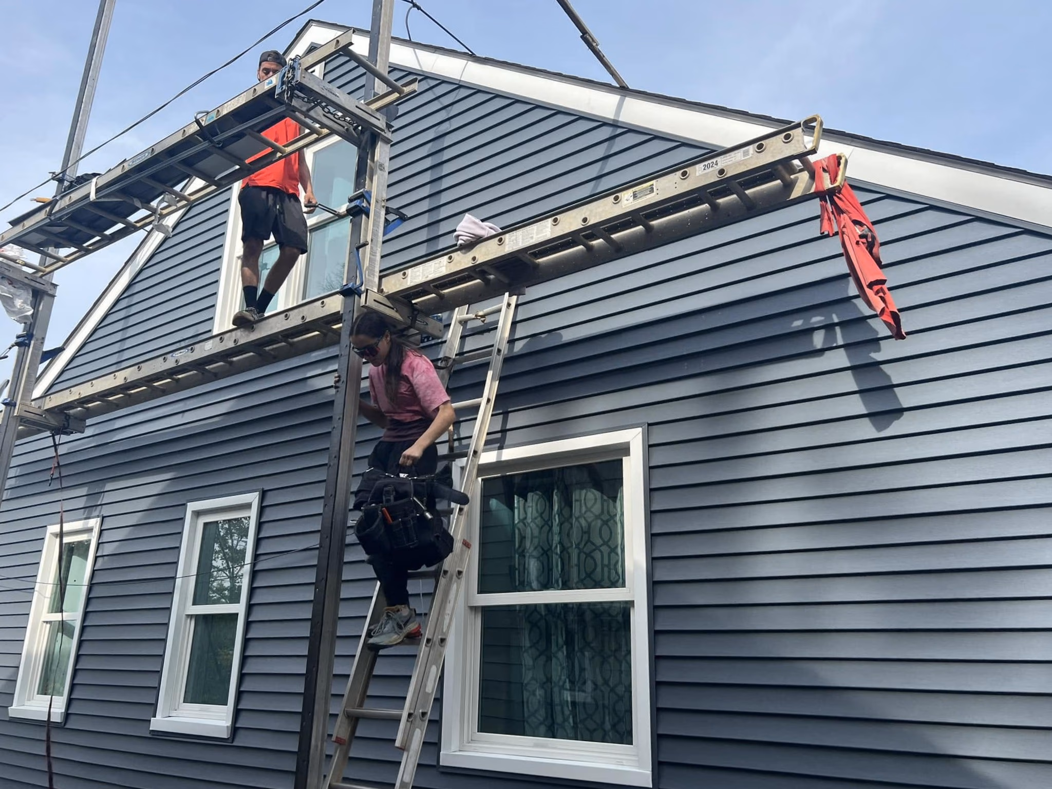 Two workers installing or repairing blue siding on a house using scaffolding and a ladder.