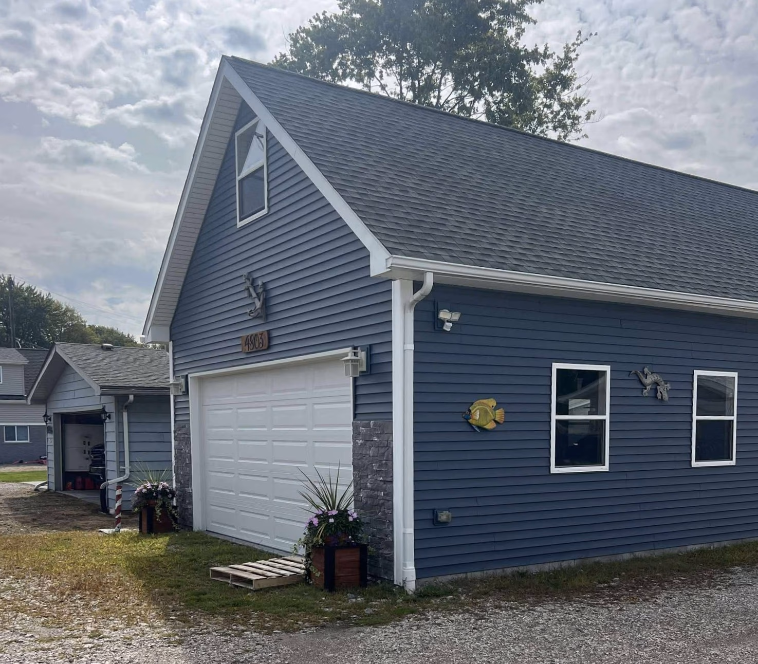 Blue garage building with white trim, a closed door, decorative fish, and plants by the entrance.