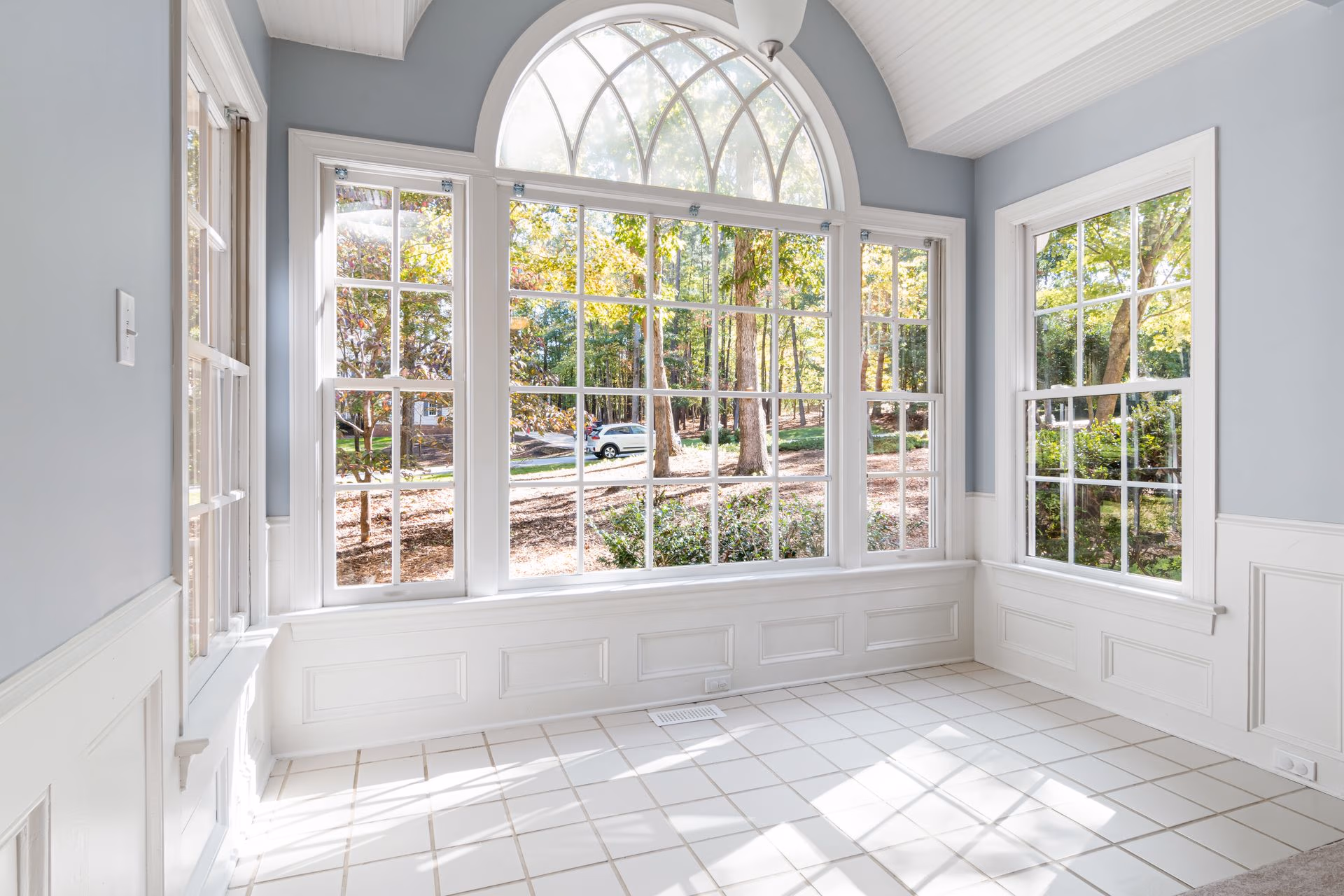 Bright empty room with large white-framed windows showing trees and a parked car outside.