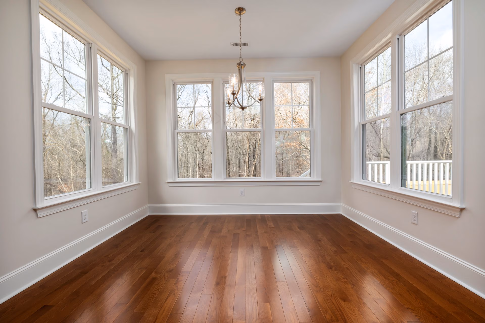 Empty room with hardwood floors, beige walls, multiple large windows, and a central ceiling light fixture.